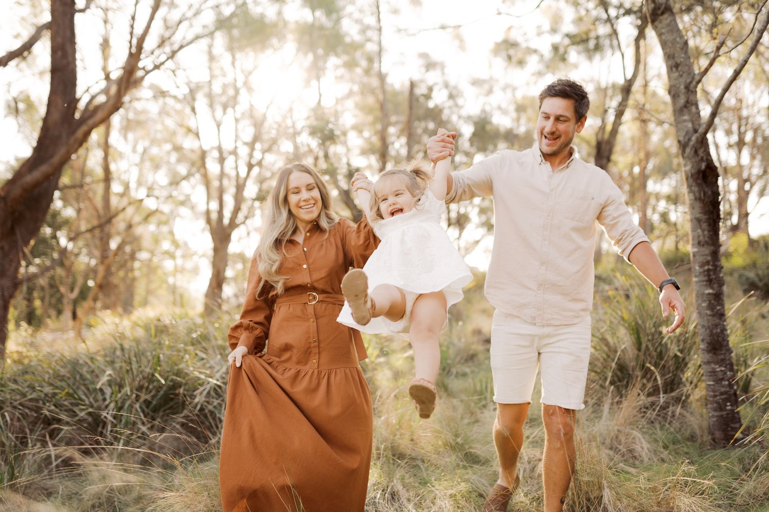 Little toddler laughing while being swung between Mother and Father in the forest during golden hour.