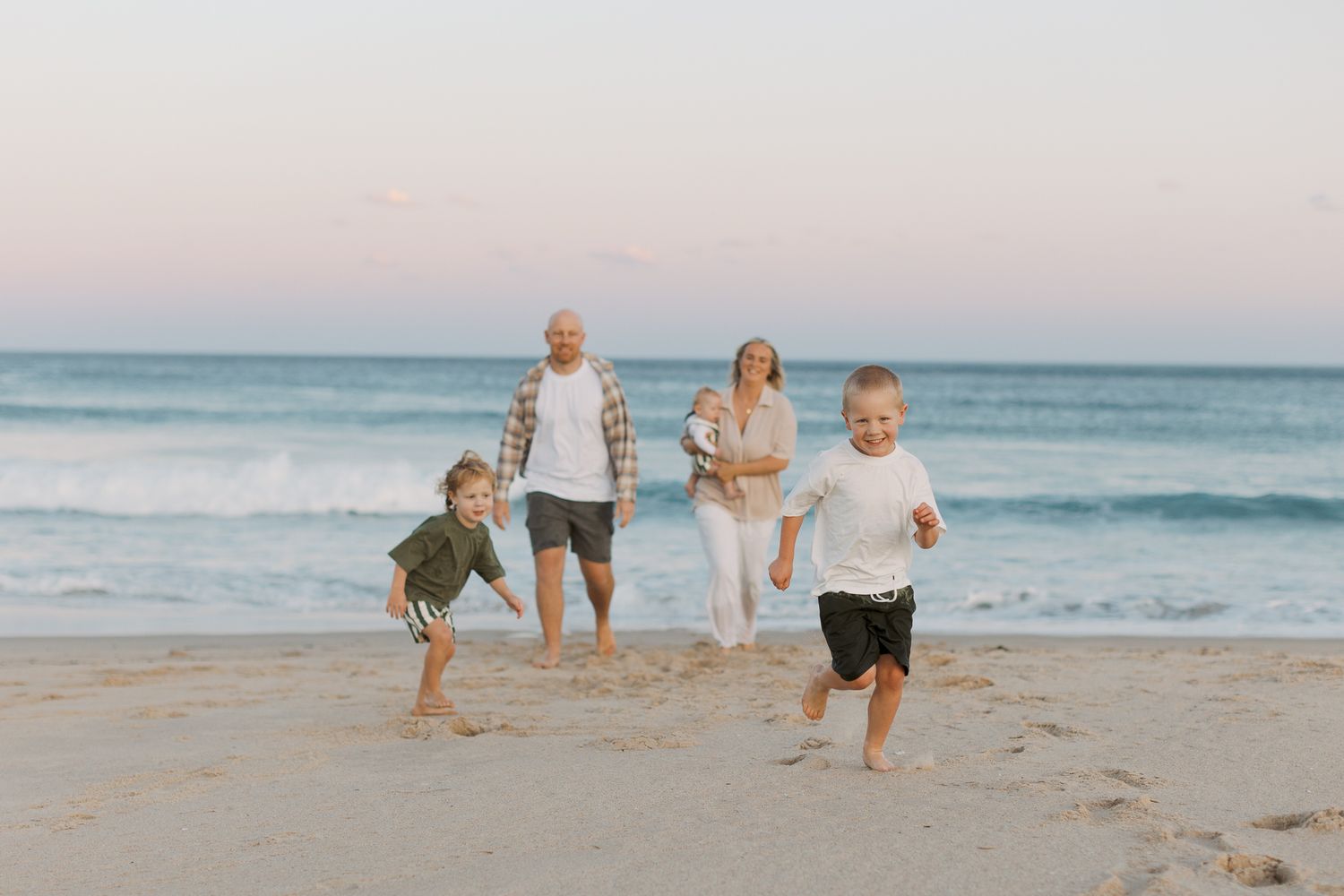 Two young boyrs running towards the camera while Mum and Dad walk slowly behind them with their youngest at Wollongong Beach during sunset.