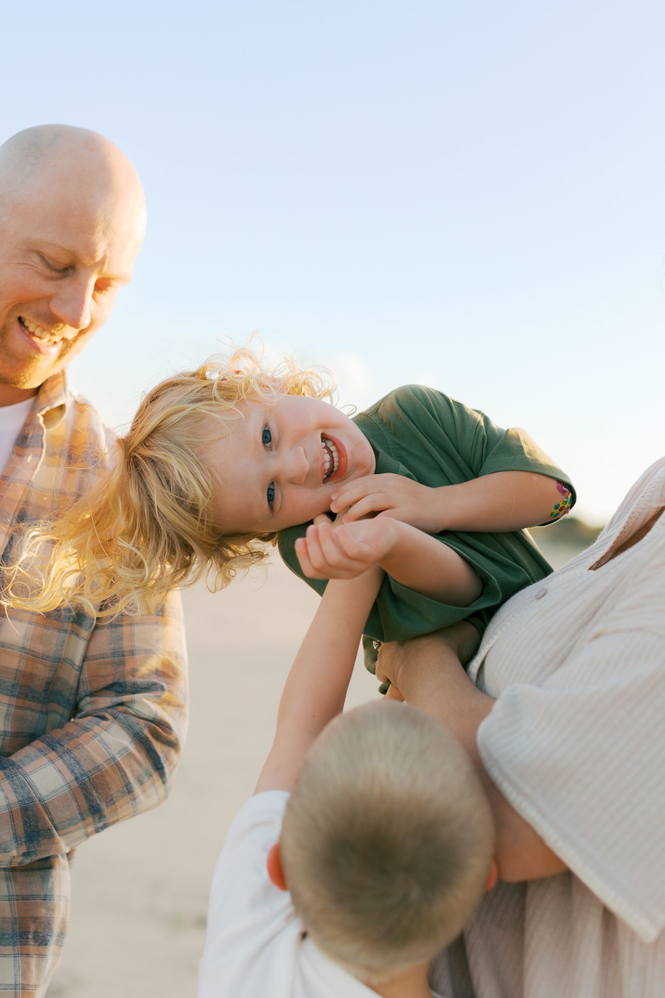 Happy young boy being held by his mother and tickled by sibling while laughing and looking directly at the camera