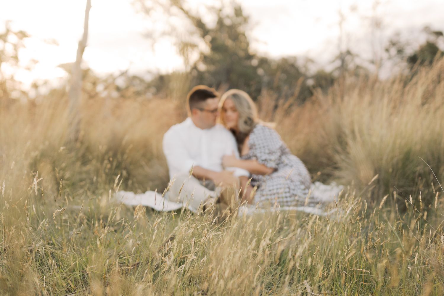 Couple sitting together and cuddling in the long grass of a summer field during golden hour.