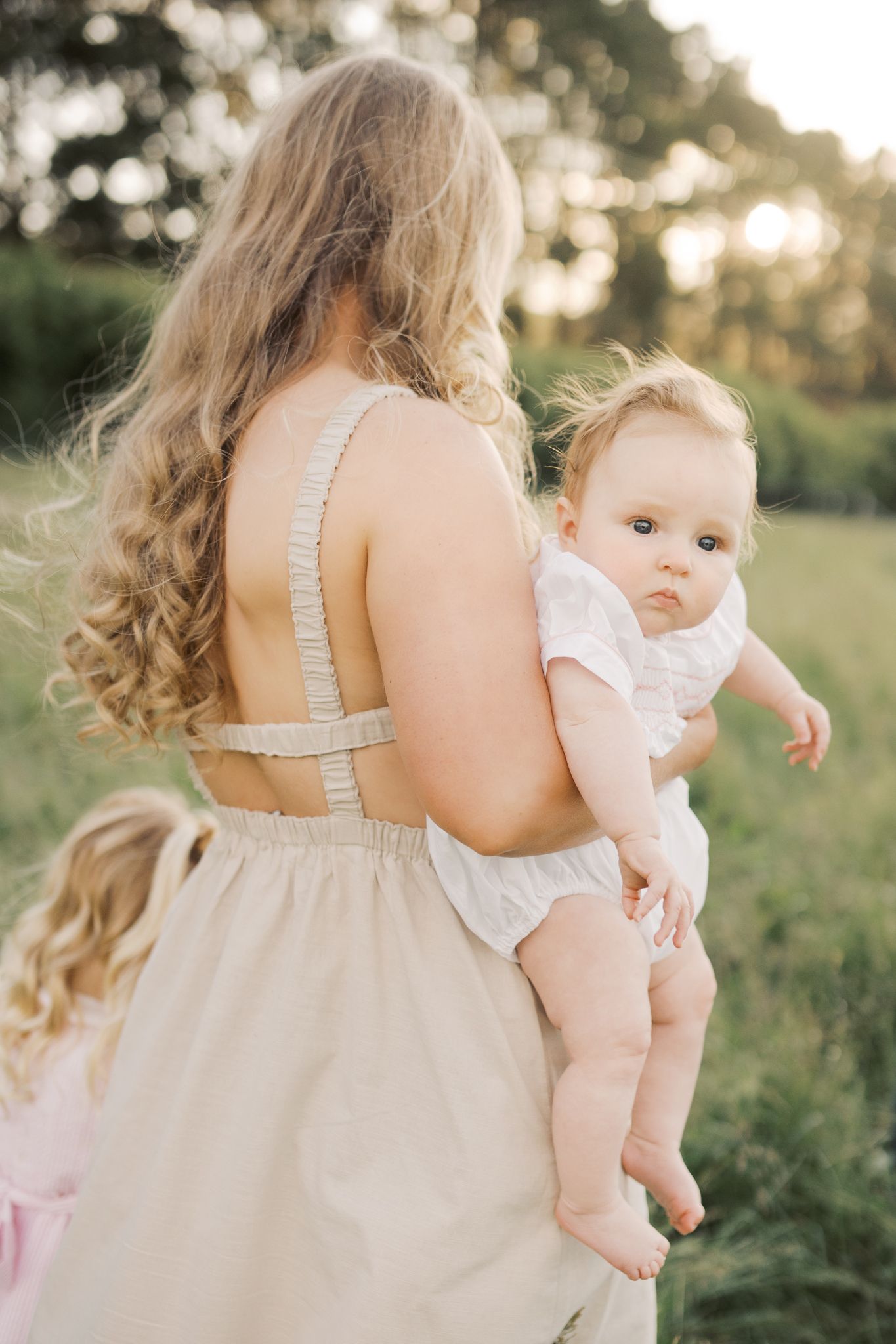 Baby girl looking back towards camera while being held by her mother as she walks away in a firld of green grass at golden hour.