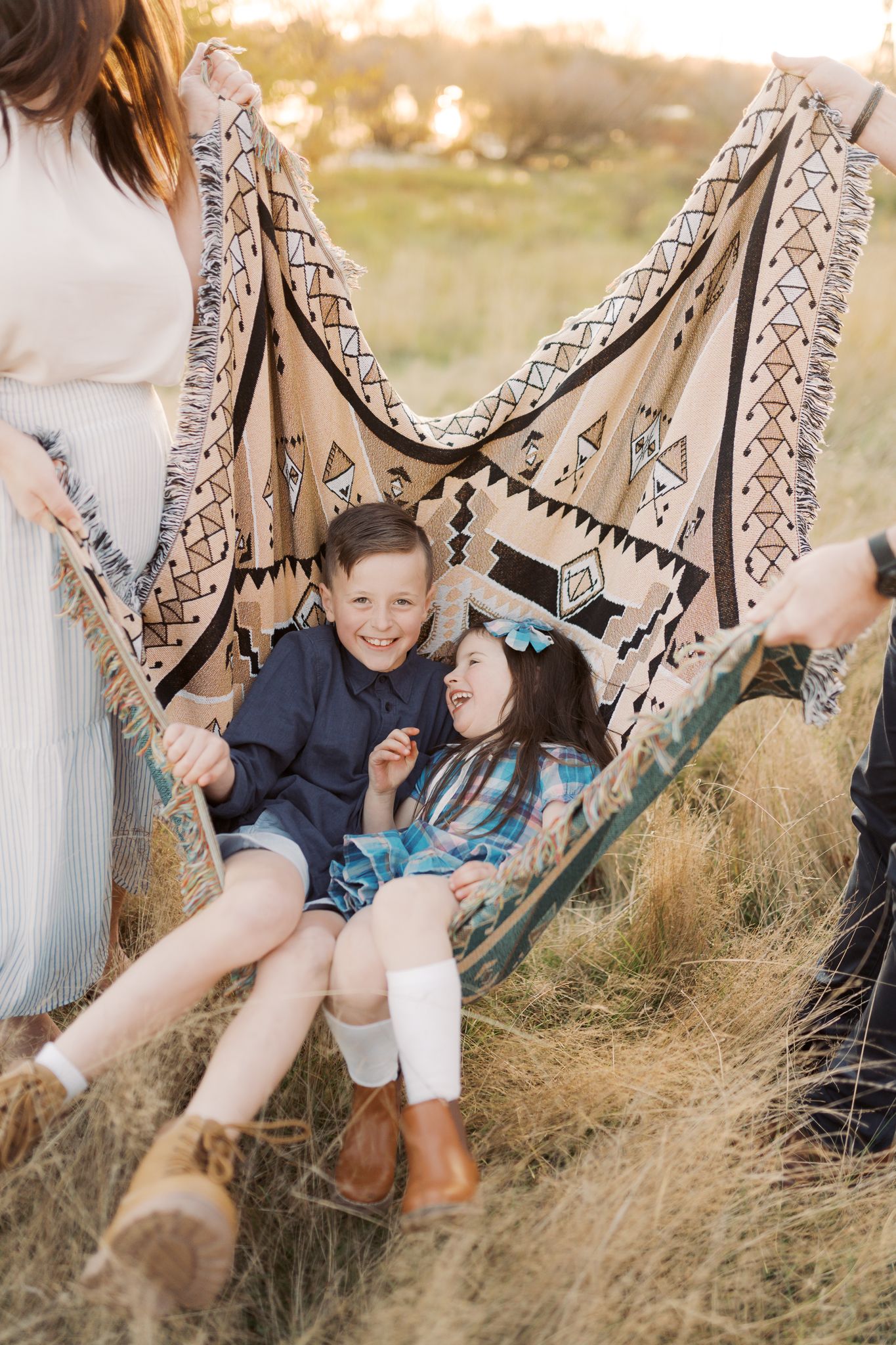 Two young siblings laughing and sitting in a picnic blanket that is turned into a hammock while Mum and Dad hold each side of the blanket.
