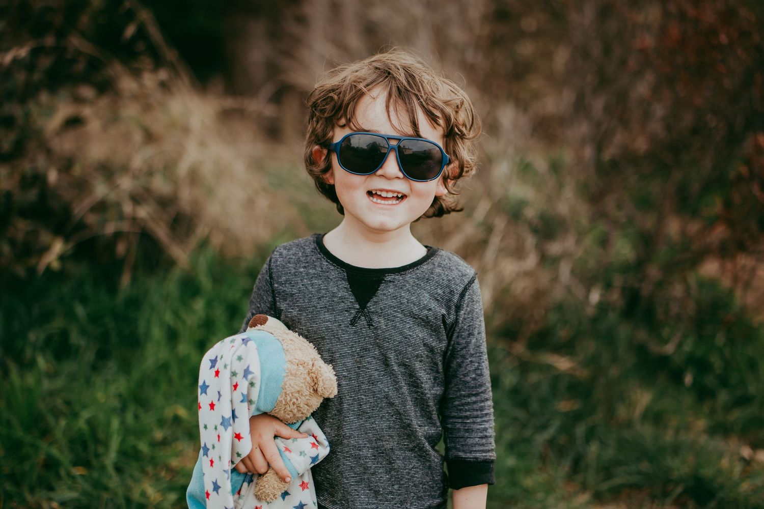 Toddler boy standing in a backyard smiling with long brown hair, wearing oversized sunglasses and holding his teddy bear.