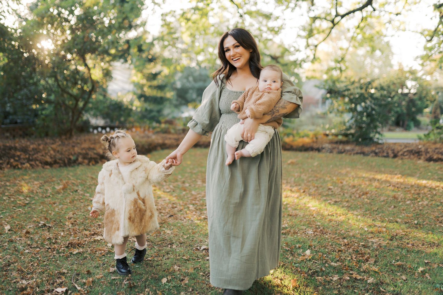 Mother smiling and holding her baby while also holding her toddler's hand walking through autumn garden at golden hour.