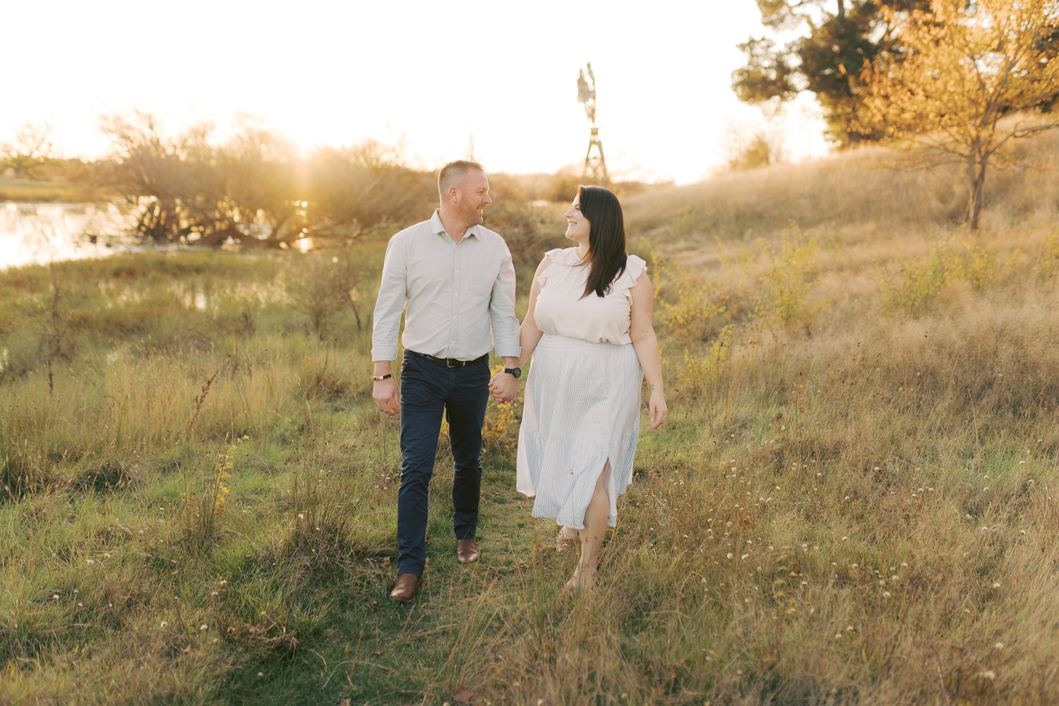 Mum and dad holding hands walking through a picturesque grassy field during golden hour with a windmill in the background