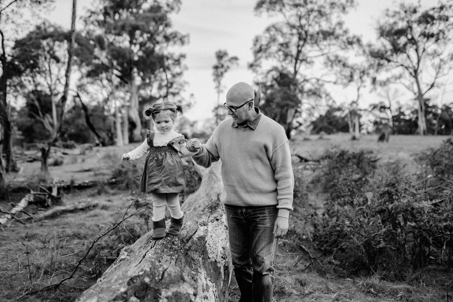Toddler balancing on log while father holds her hand.