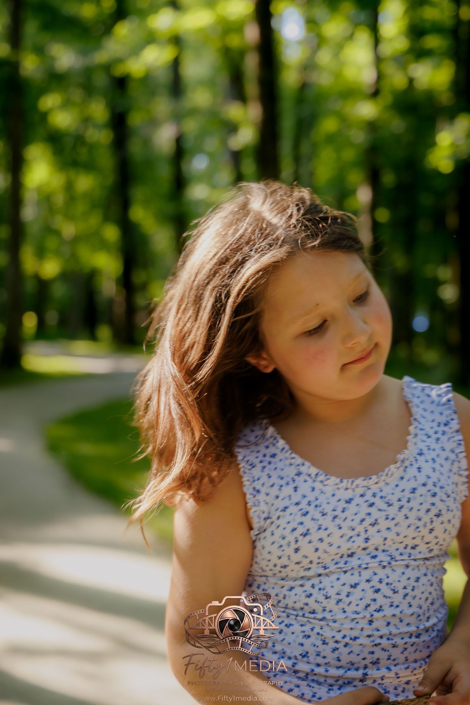 Outdoor lifestyle portrait of a young girl in a blue dress standing in a sunlit green park.