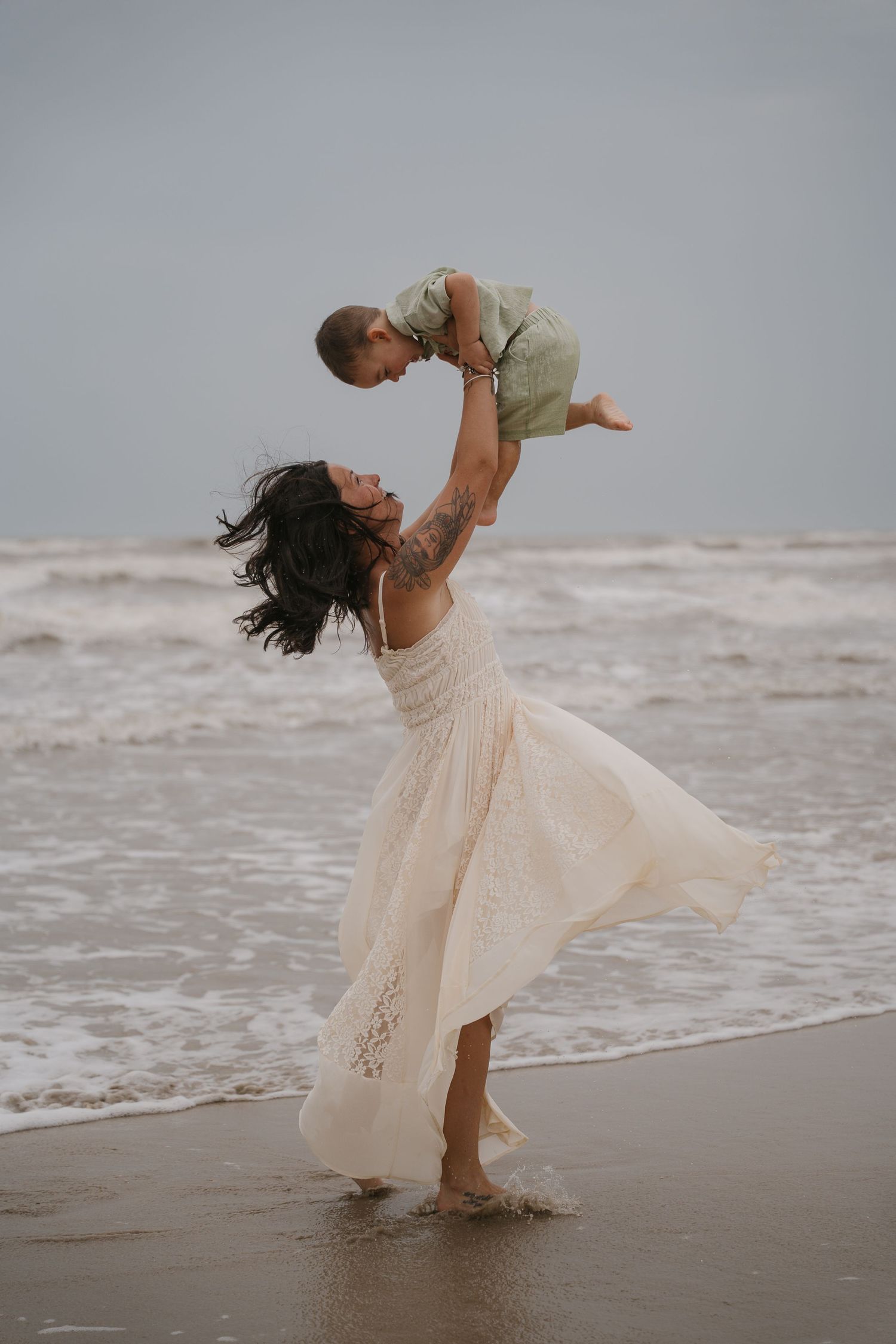 Woman in white dress lifting baby overhead on beach with ocean waves, joyful mother-child moment.