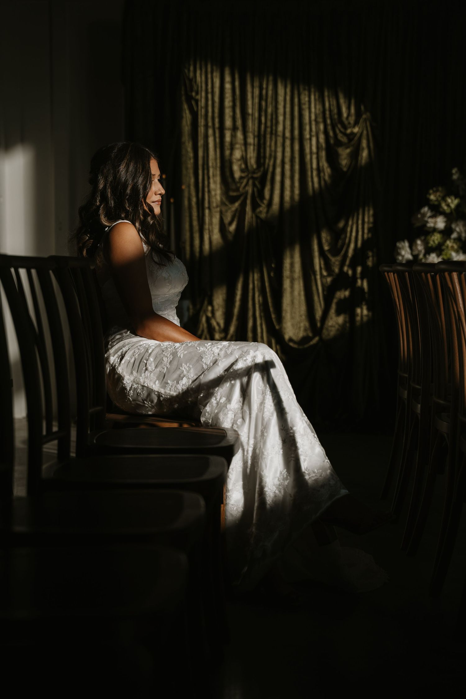 Bride in white lace gown sits peacefully in dramatic window light inside a darkened room.