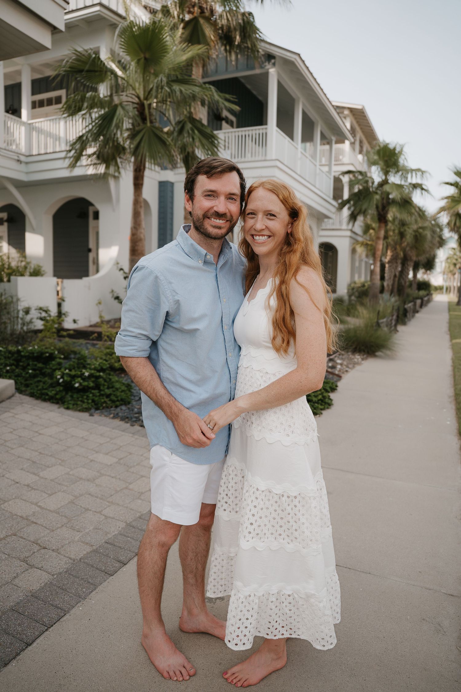 Smiling couple in casual beach attire posing barefoot on a sidewalk with palm trees and coastal homes behind them.