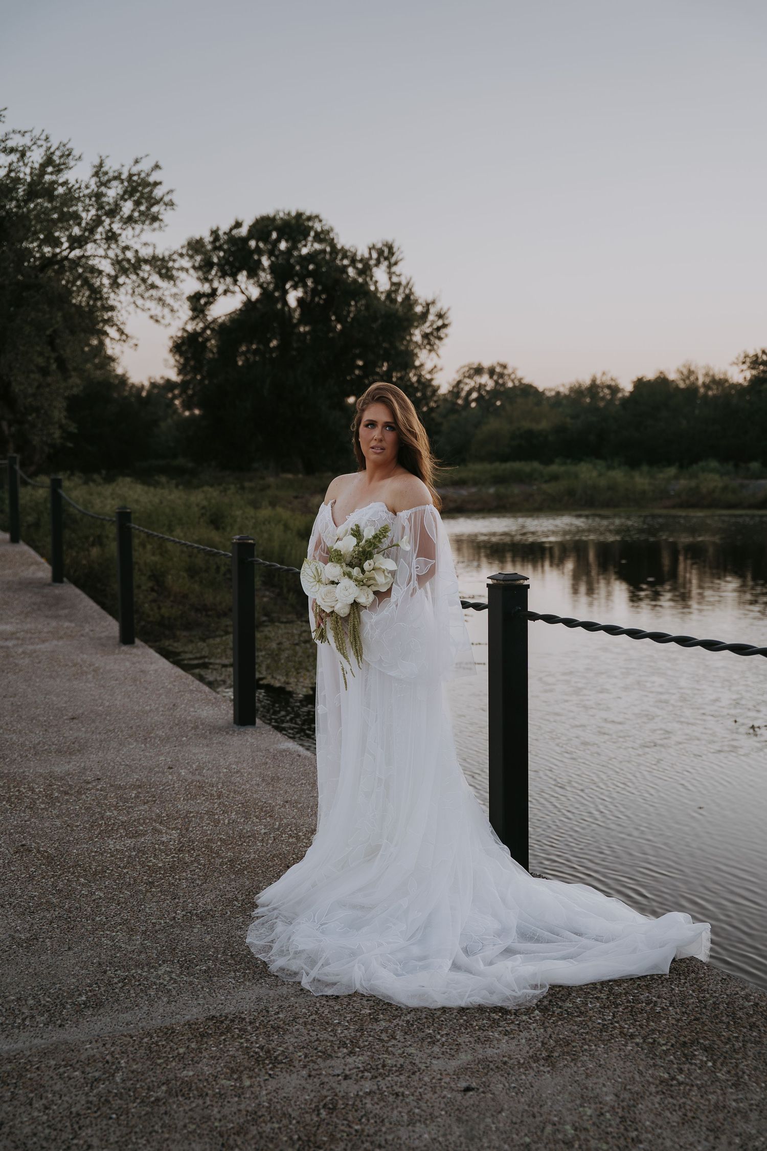 Bride in flowing white off-shoulder gown holding bouquet stands on waterfront path at sunset.