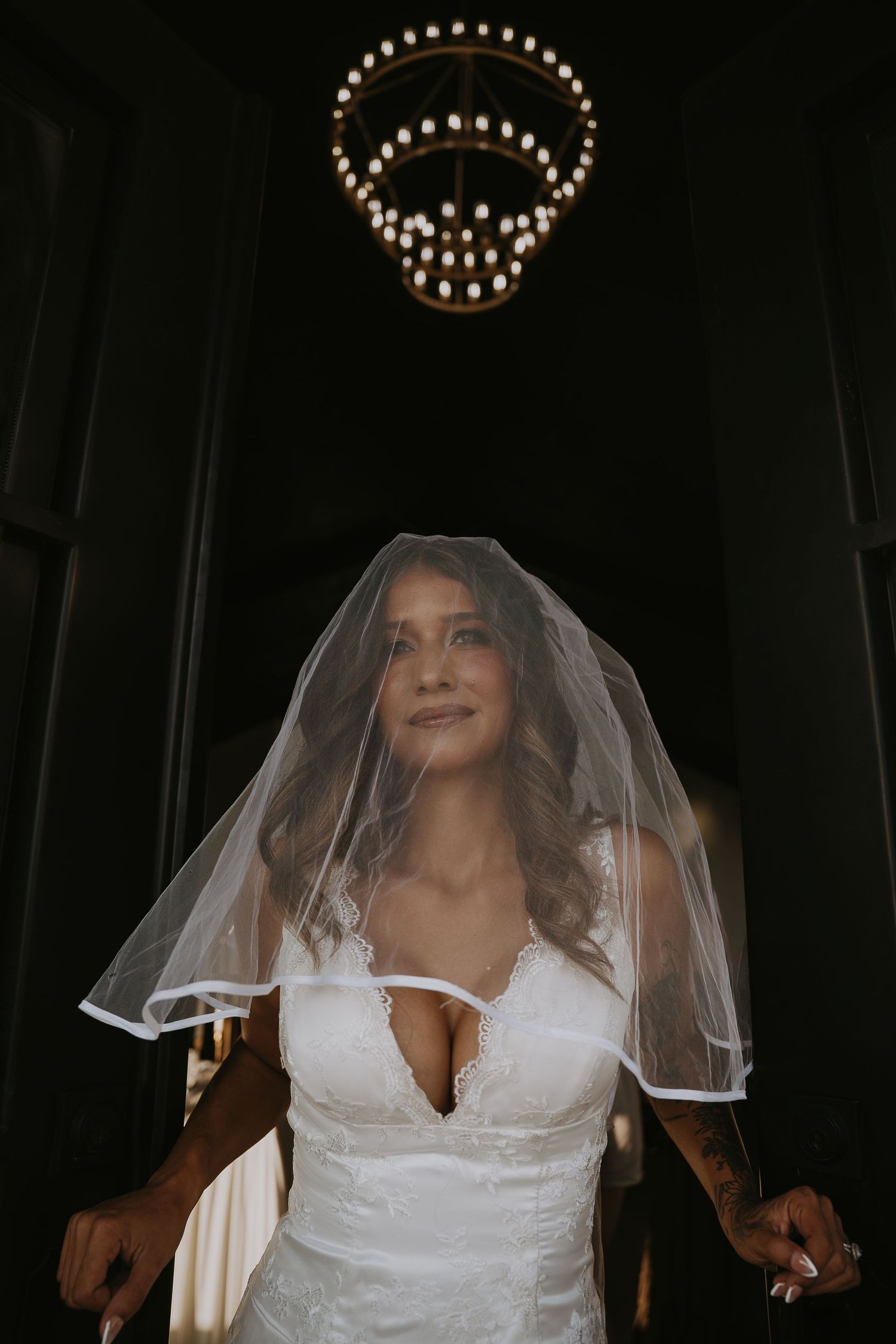 Beautiful bride in lace wedding dress and veil stands beneath elegant chandelier in dark venue.