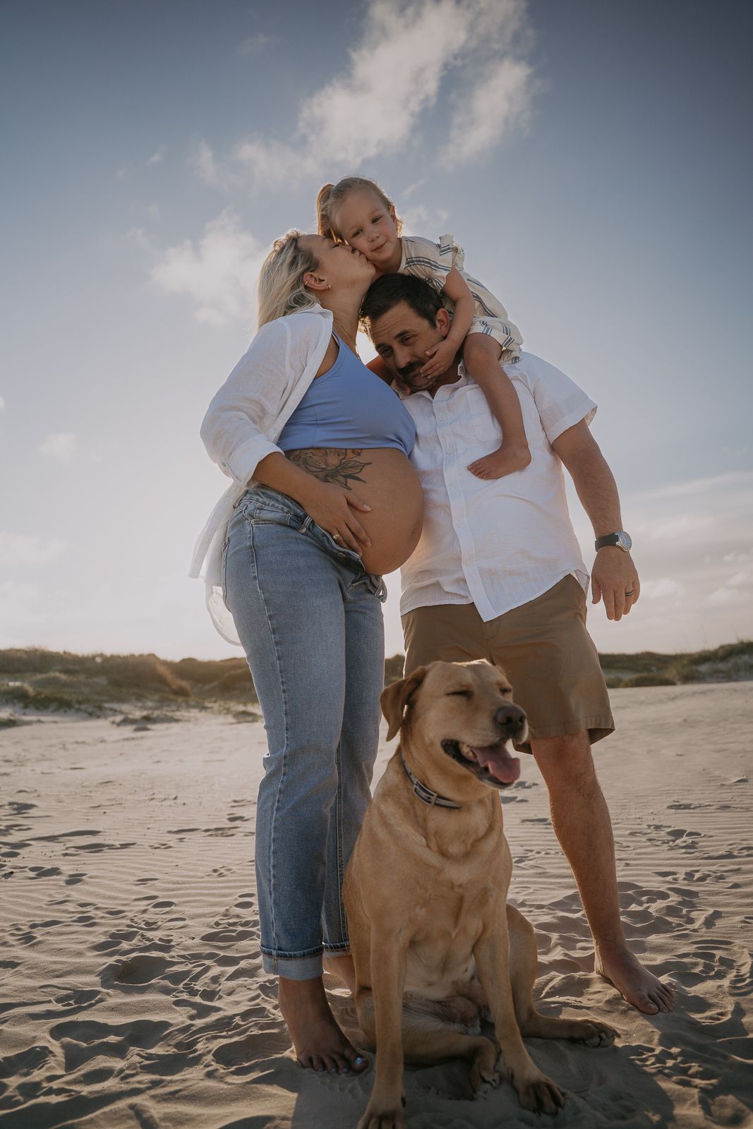 Pregnant family with young child and Labrador dog enjoy golden hour beach portrait together.