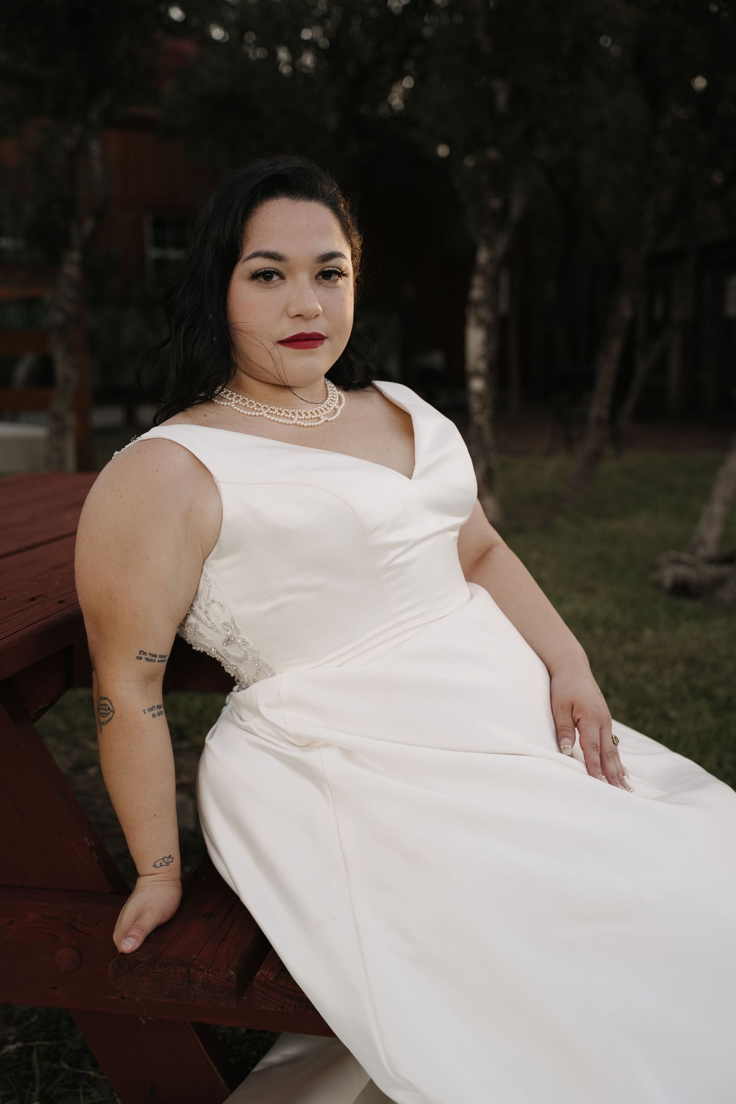 Beautiful bride in white satin gown with pearl necklace poses on red bench in garden setting.
