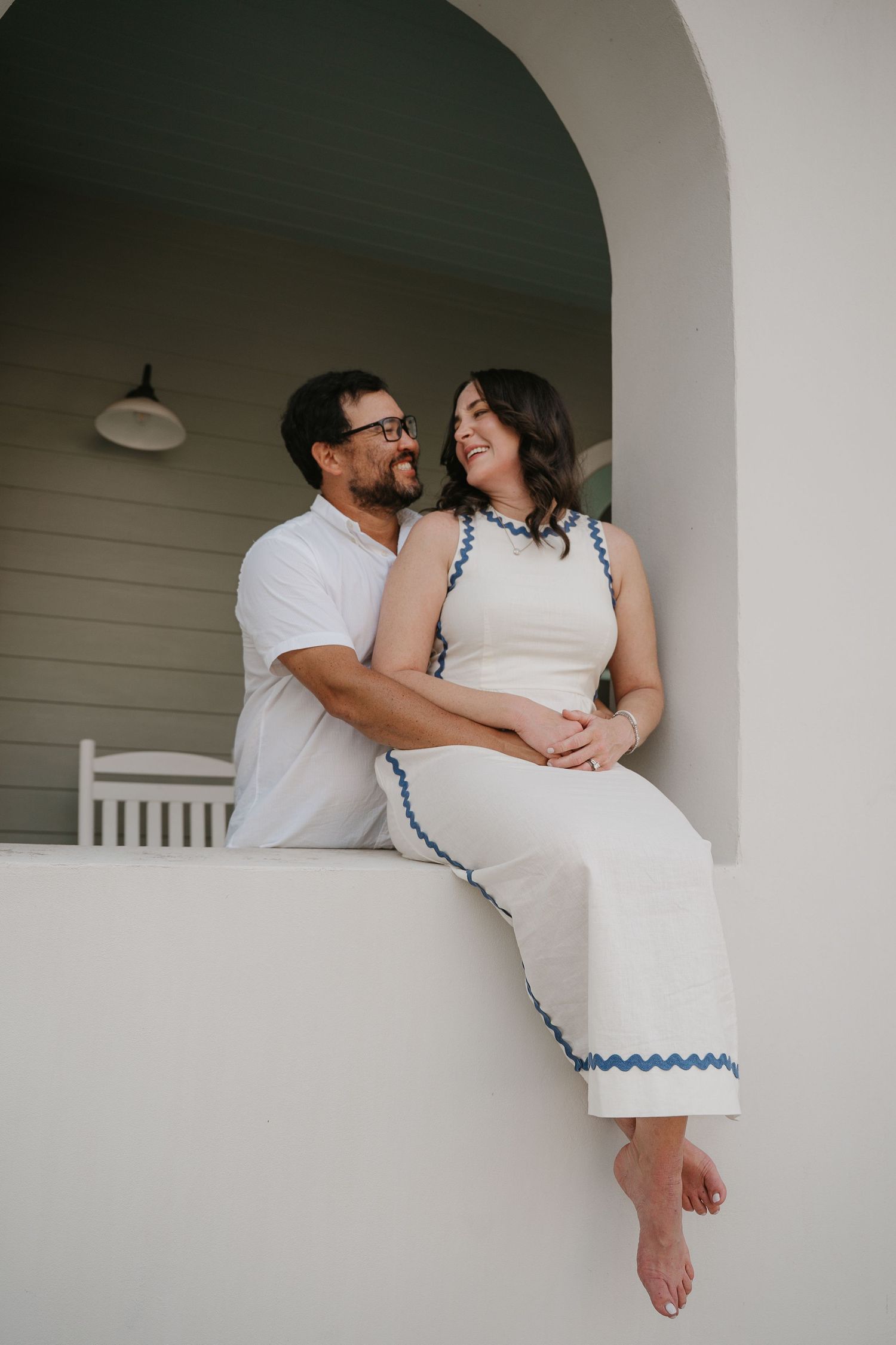 A couple shares a loving glance on a white porch ledge, woman in white dress with blue trim, man in white shirt.