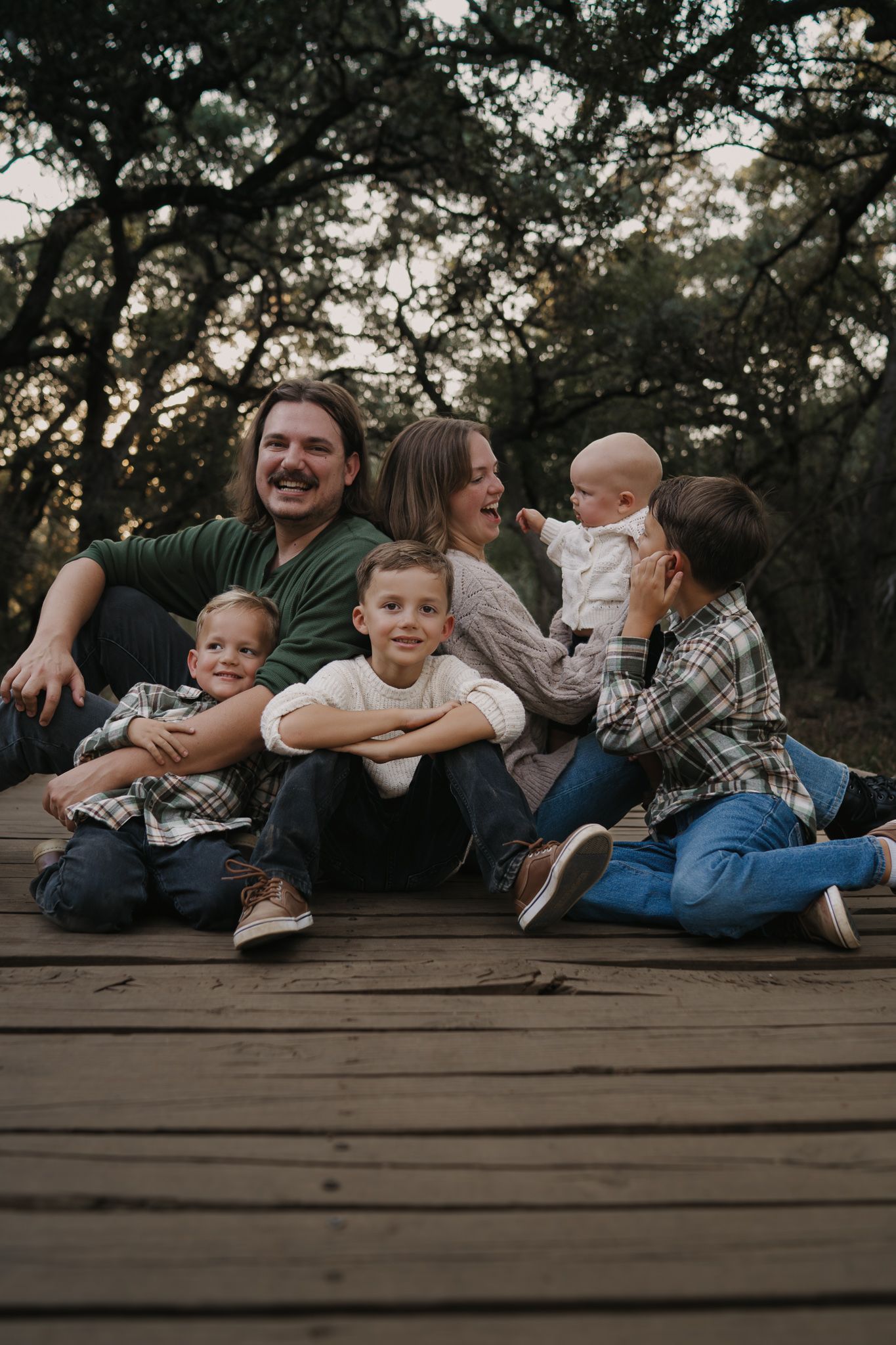 Happy family of six sitting together laughing on wooden deck surrounded by trees.