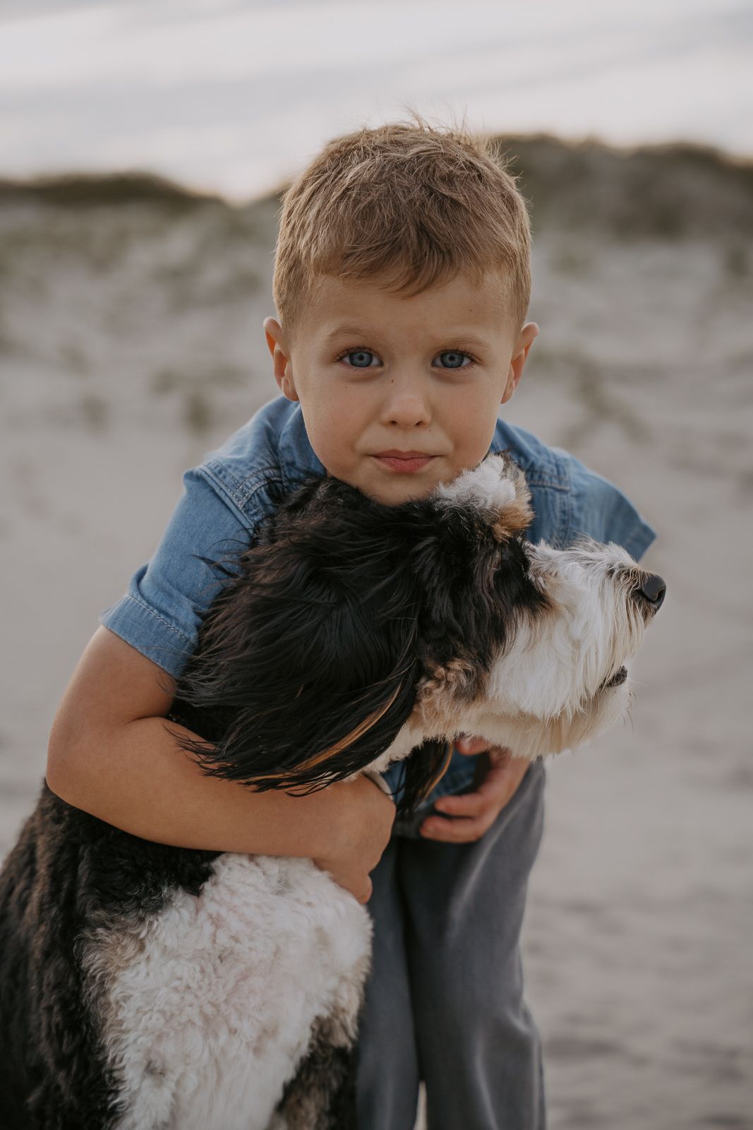 Adorable blonde boy with blue eyes hugging a black and white dog on the beach.
