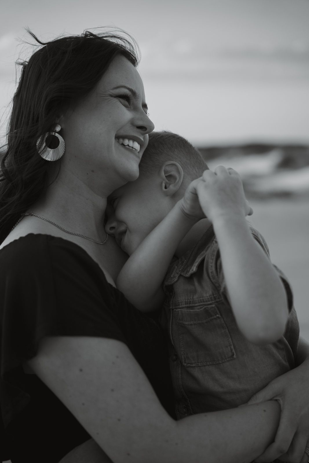 Black and white portrait of laughing mother hugging young child at the beach.