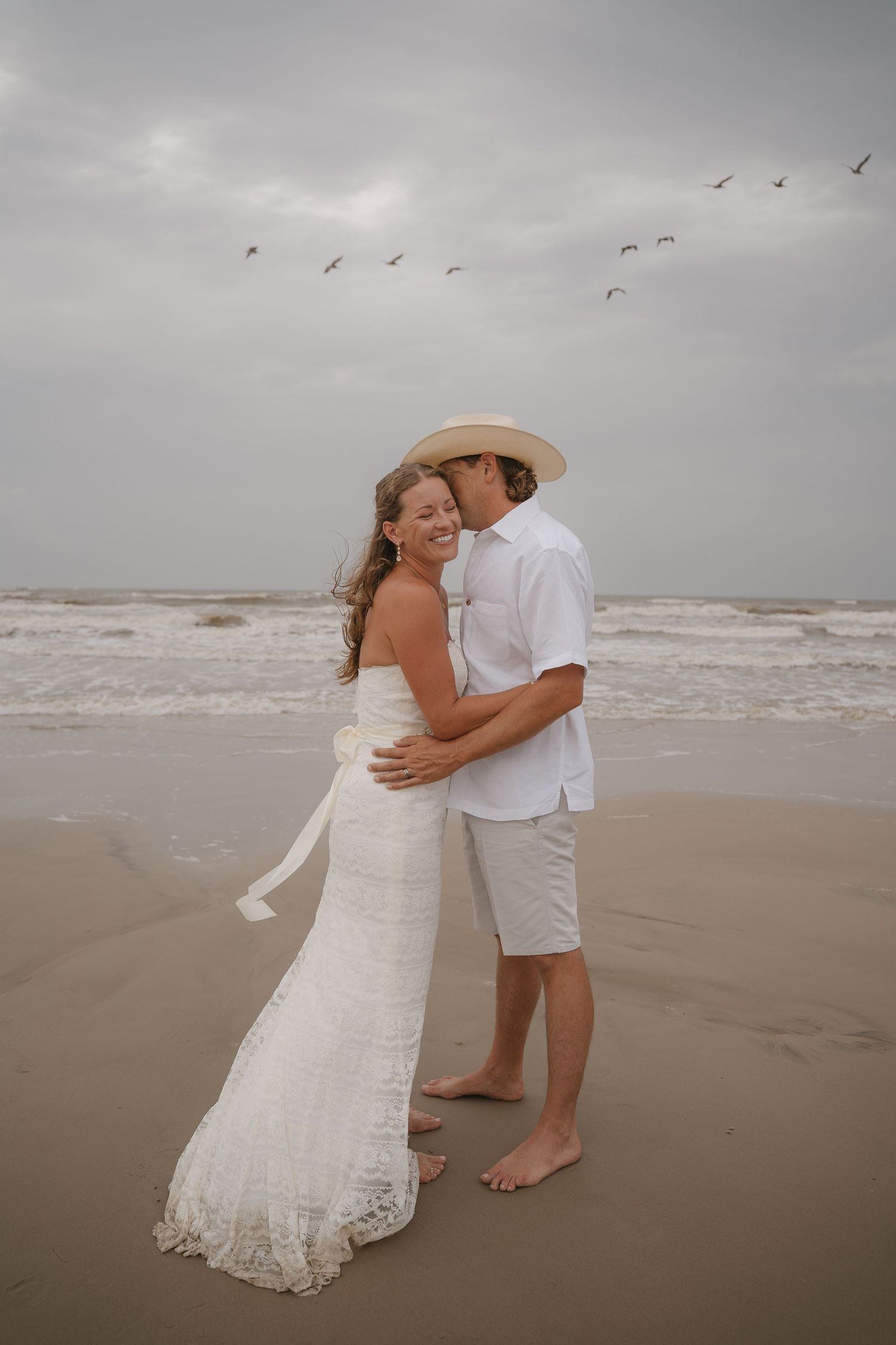 Couple in white embracing on a beach, bride in lace dress and groom in hat, with ocean waves and birds in cloudy sky.