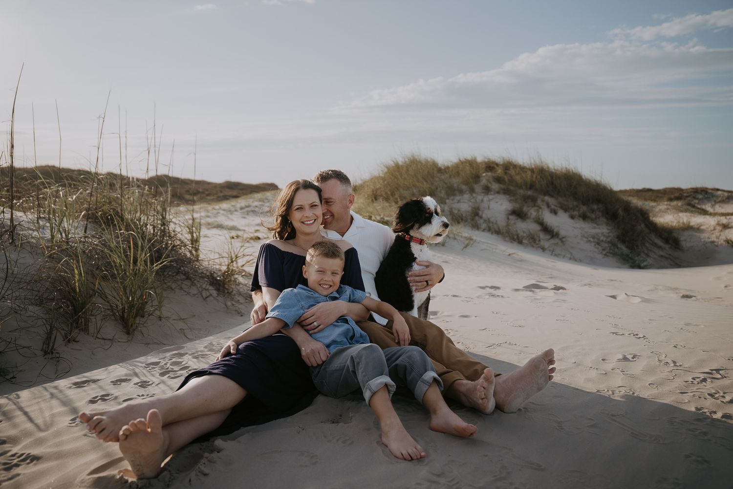 Happy family with dog sitting together on sandy beach dunes.