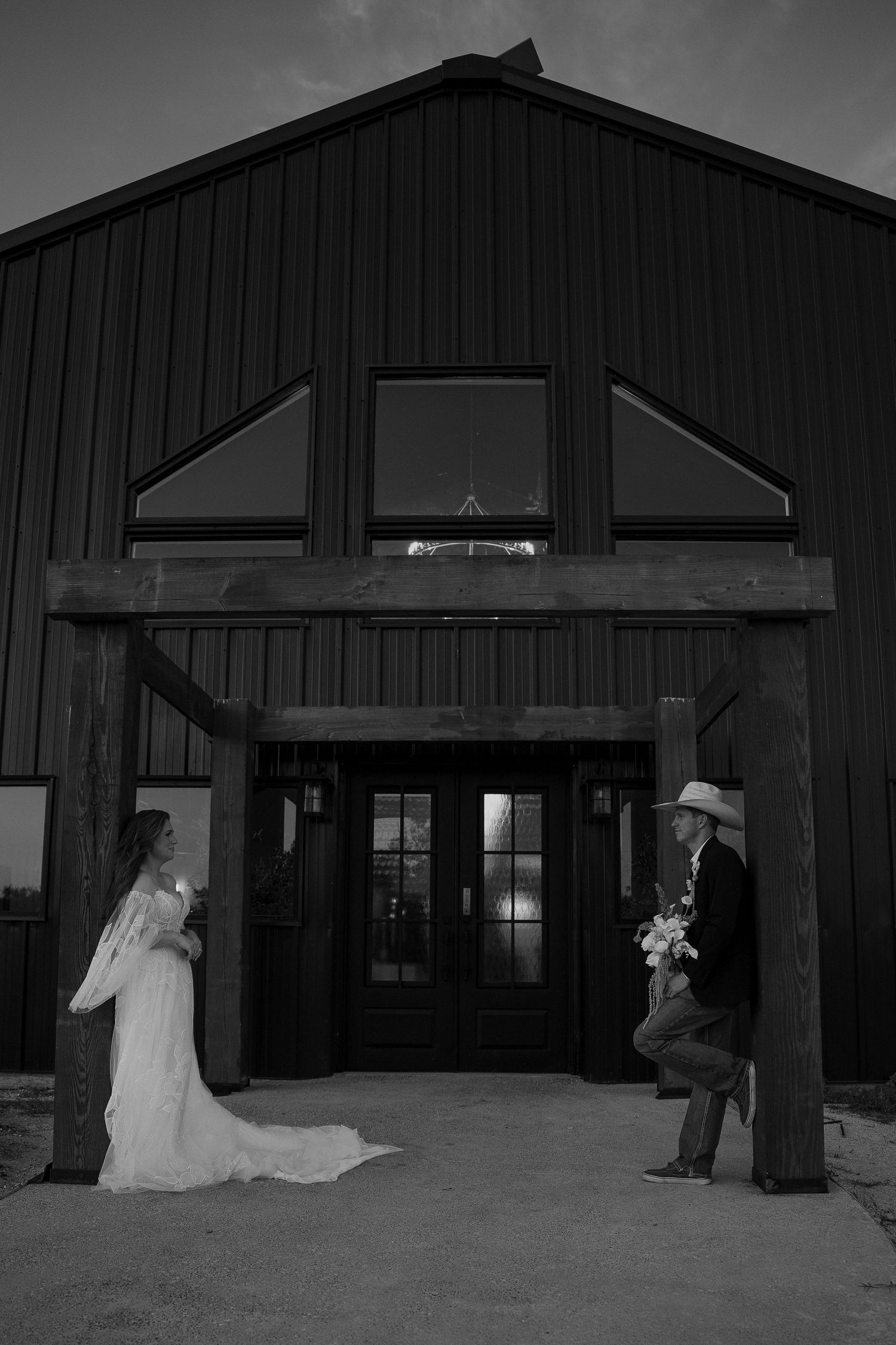 Black and white photo of bride and groom in cowboy hat outside dark barn at dusk.