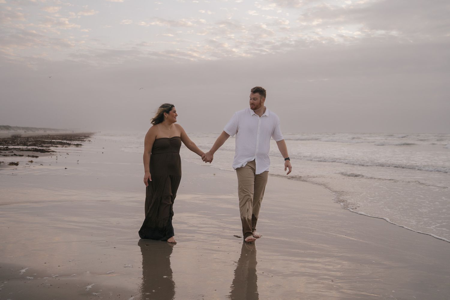 Couple holds hands walking barefoot along ocean shoreline at sunset during romantic beach session.