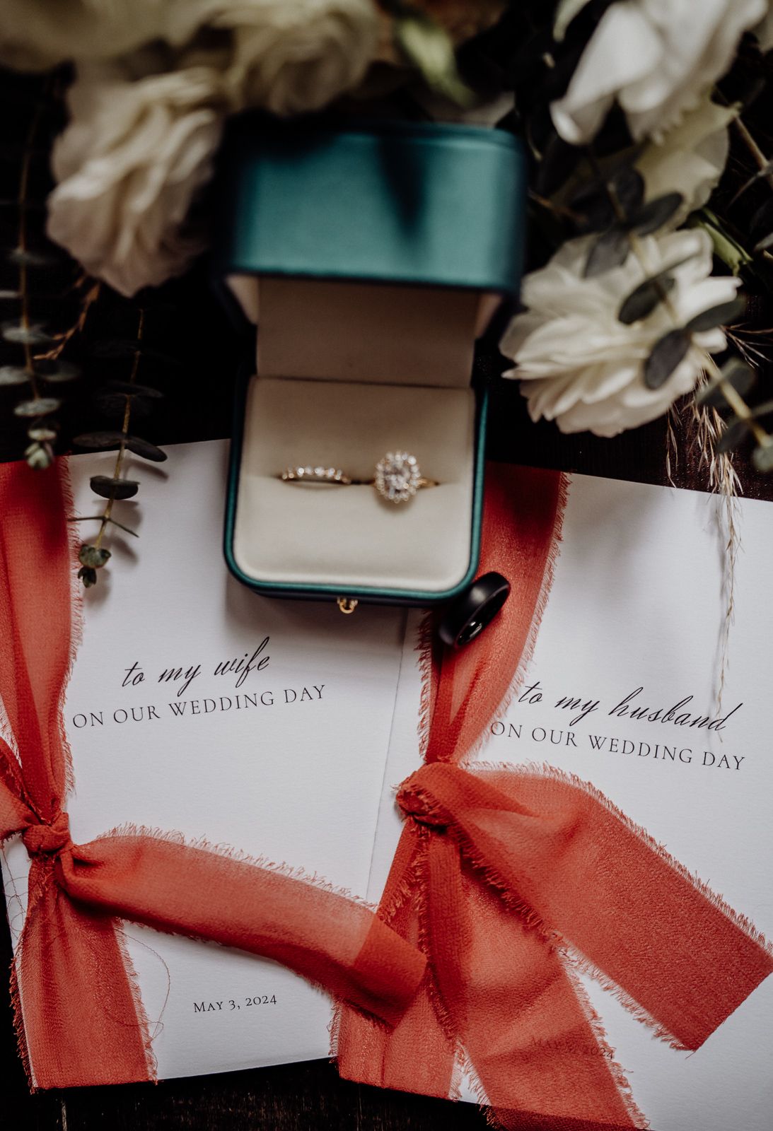 Diamond engagement ring in teal box beside vow books with red ribbons and white flowers.