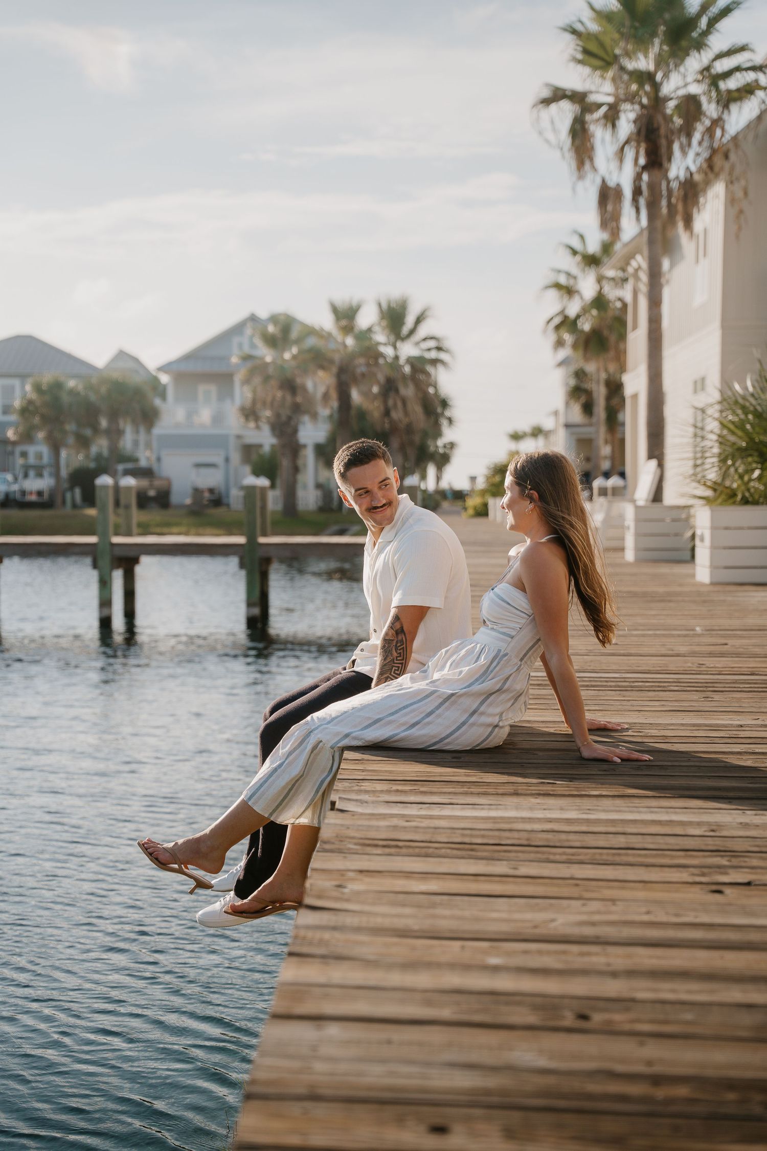 Couple sitting on a wooden dock by the water with palm trees and coastal homes in the background.