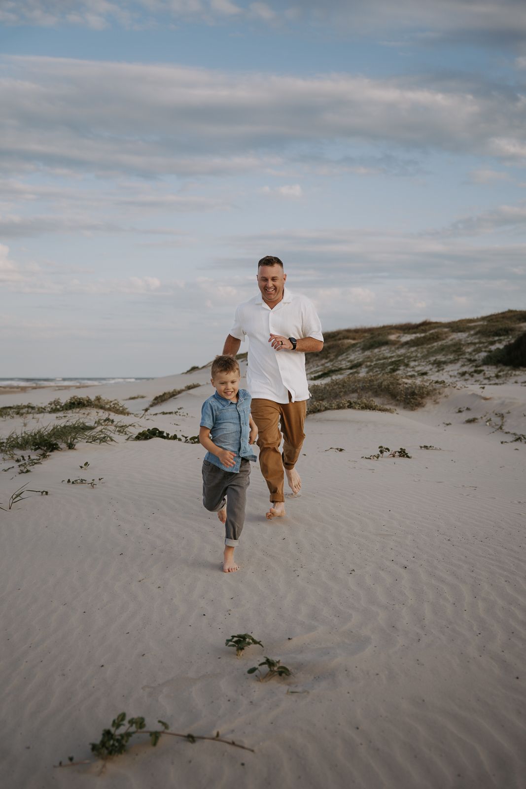 Father and young son running barefoot together on sandy beach dunes under cloudy sky.