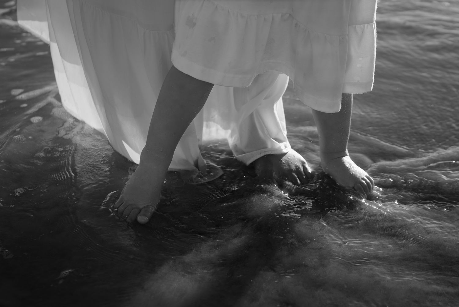 Black and white photo of barefoot mother and child standing in ocean waves.