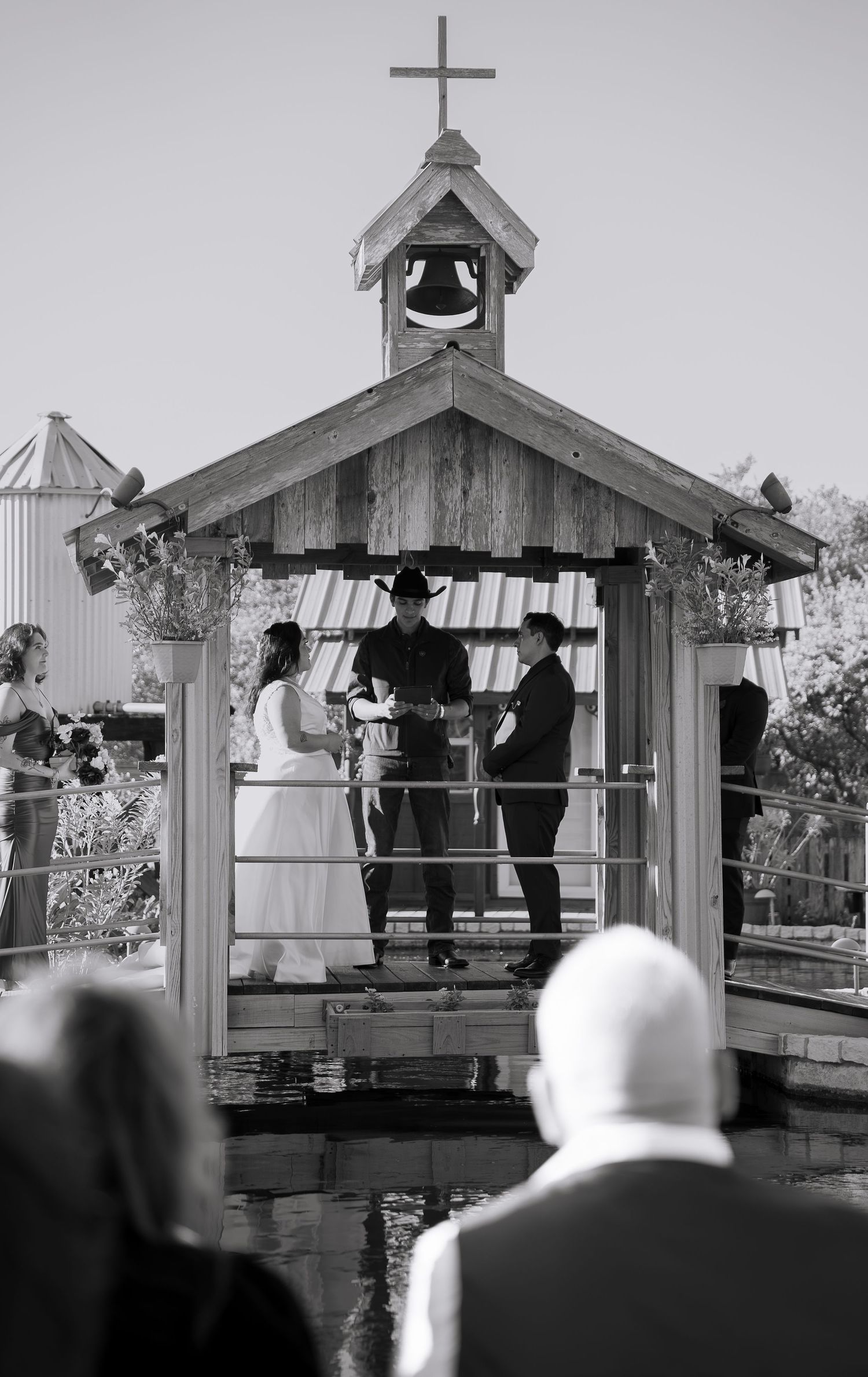 Black and white photo of a couple exchanging vows in a rustic wooden chapel over water.