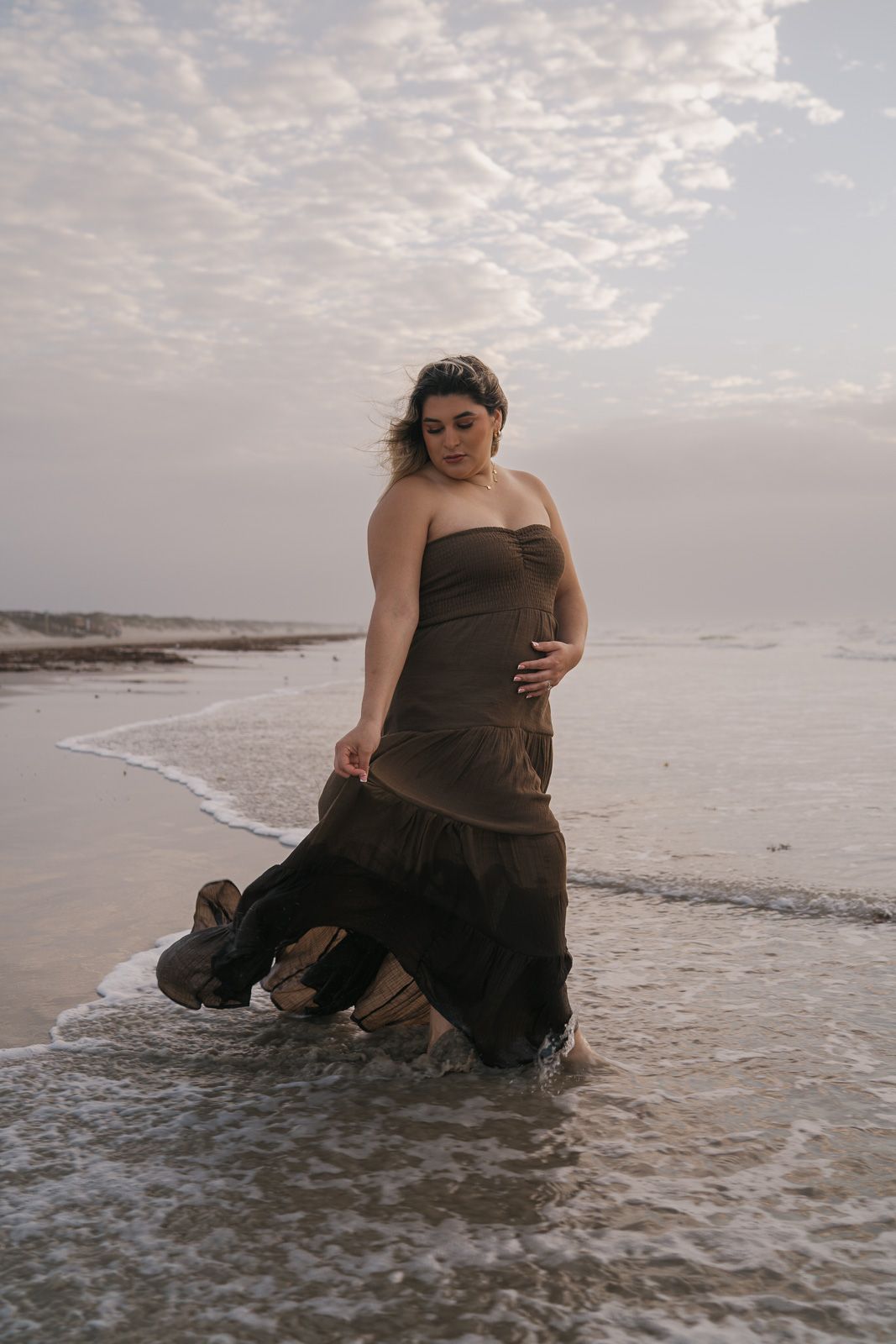 Pregnant woman in brown strapless gown standing in ocean waves on cloudy beach.