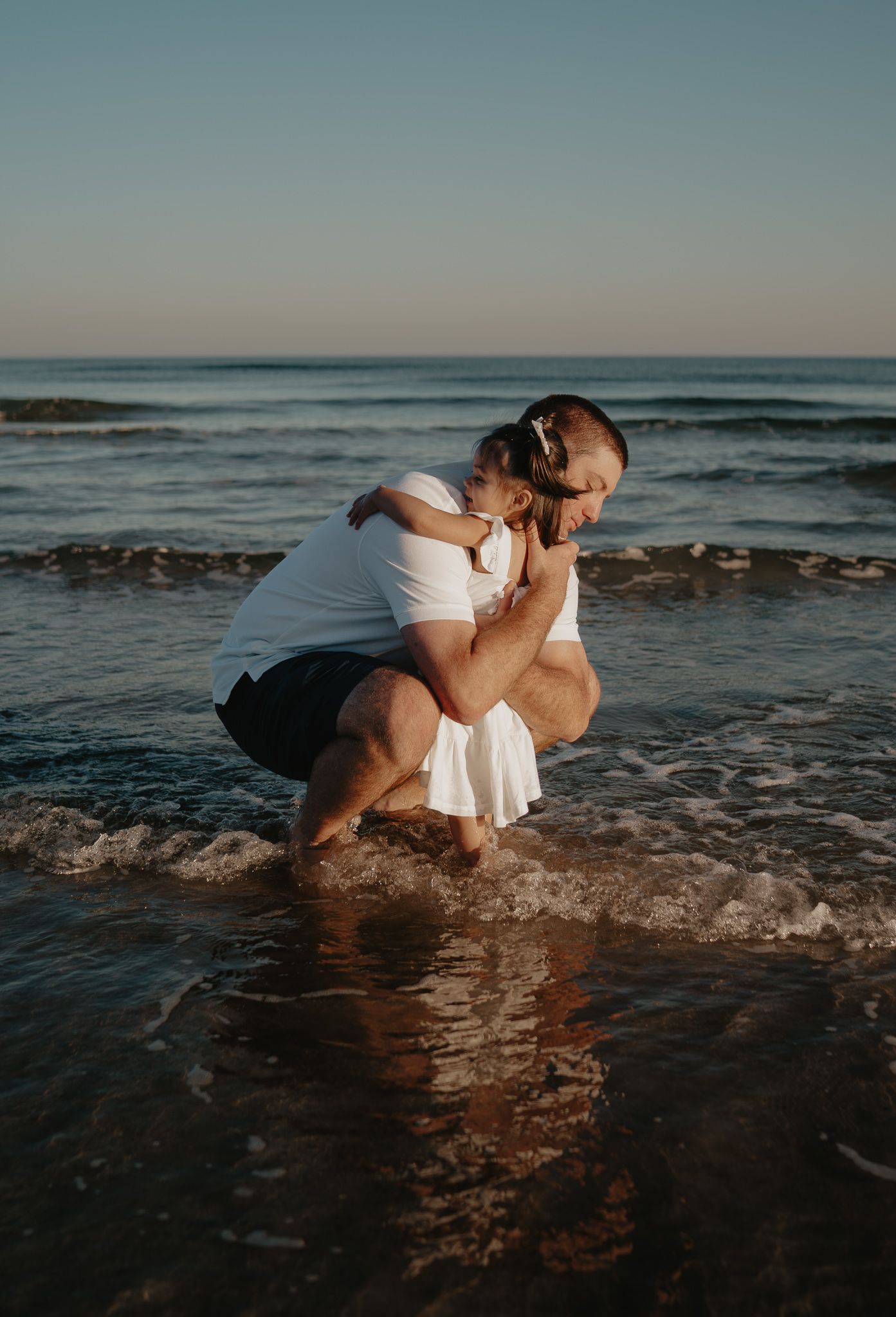 Father crouching in ocean waves tenderly holding and kissing his toddler at sunset beach.