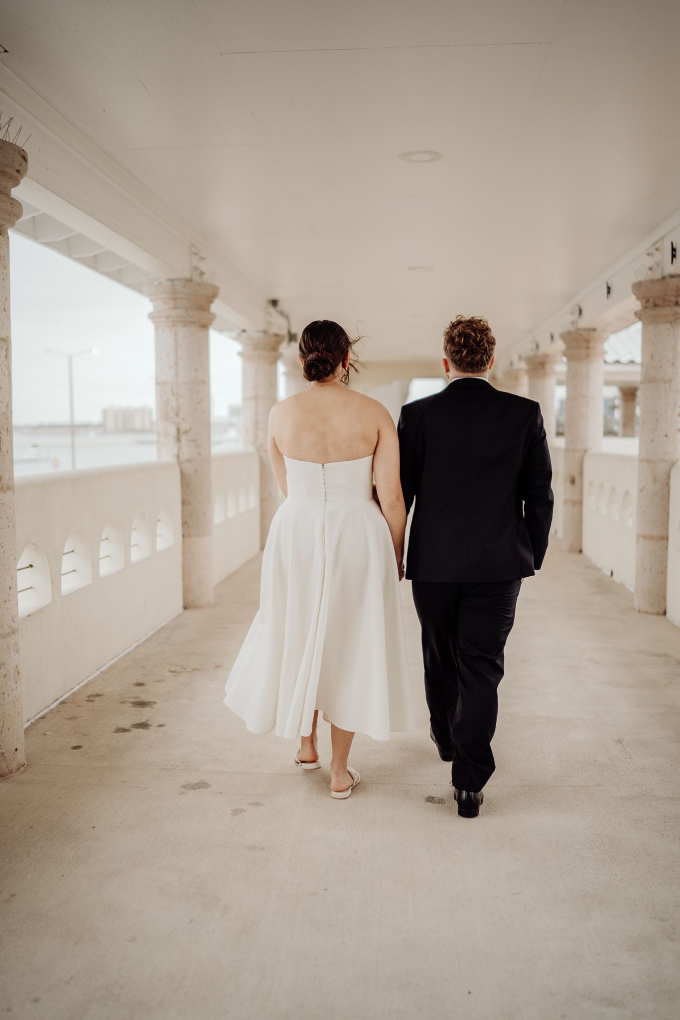 Bride in white midi dress and groom in black suit walking hand in hand down elegant corridor.
