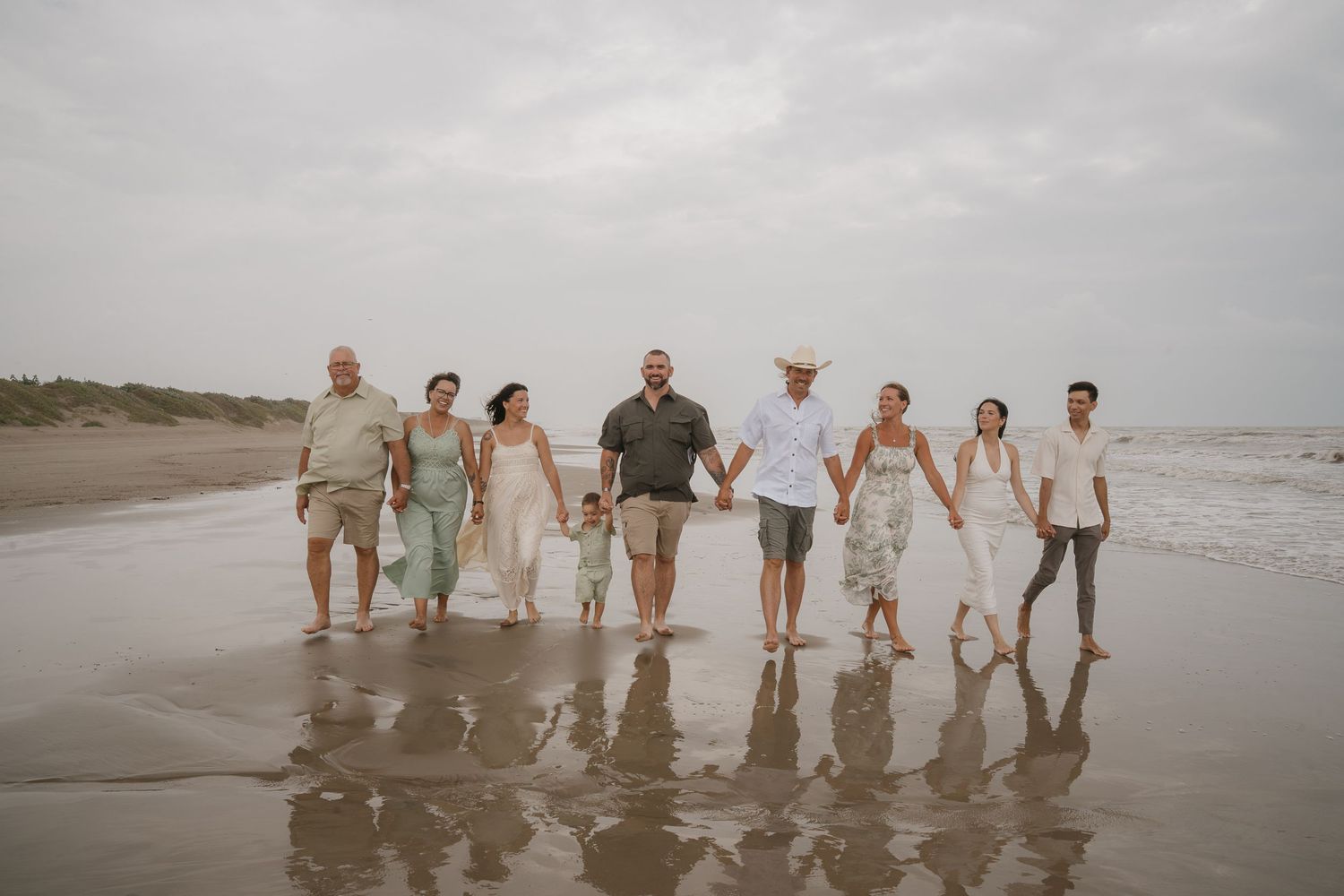 Group of eight people walking hand-in-hand on a cloudy beach, dressed in casual summer attire.