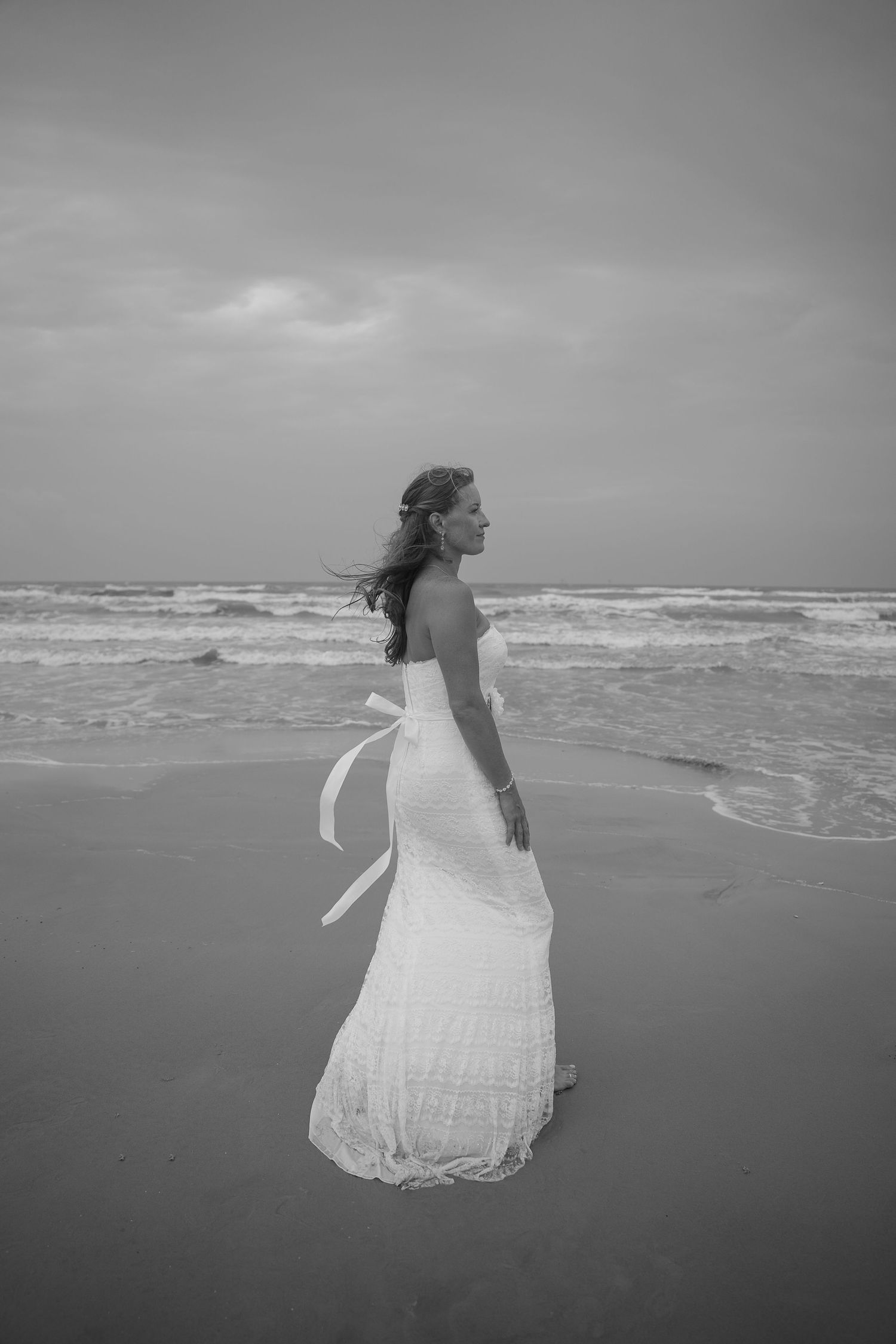 Bride in white strapless gown with ribbon bow stands on sandy beach, waves crashing behind her, black and white photo.