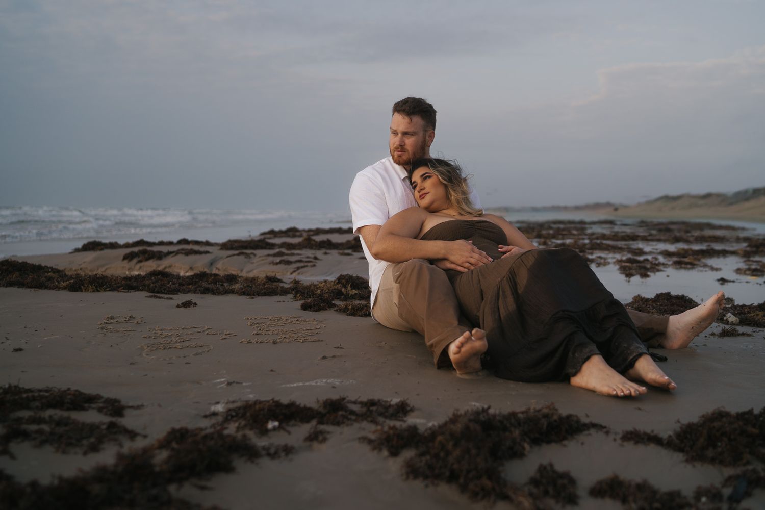 Romantic couple sitting together on sandy beach at dusk with ocean waves behind them.