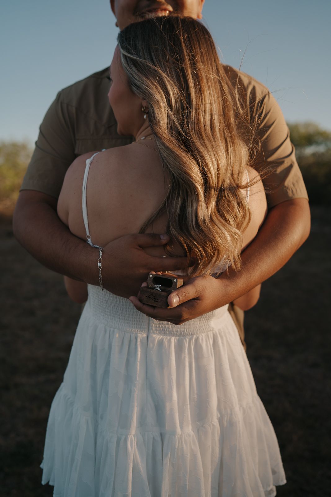Couple embracing at golden hour in an open field, woman in white skirt and spaghetti strap top during engagement session.