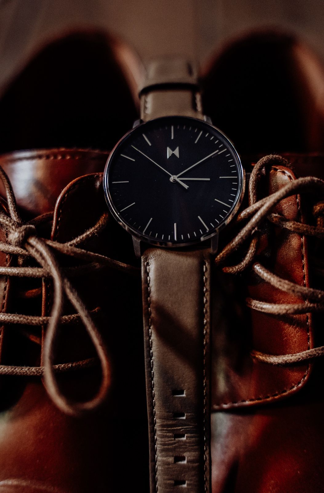 MVMT black minimalist watch resting on brown leather lace-up dress shoes for a wedding detail shot.