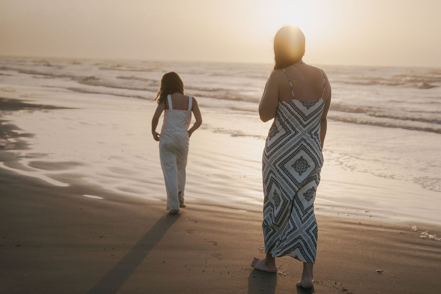 Two women in dresses walking on a sandy beach at sunset with ocean waves in the background.