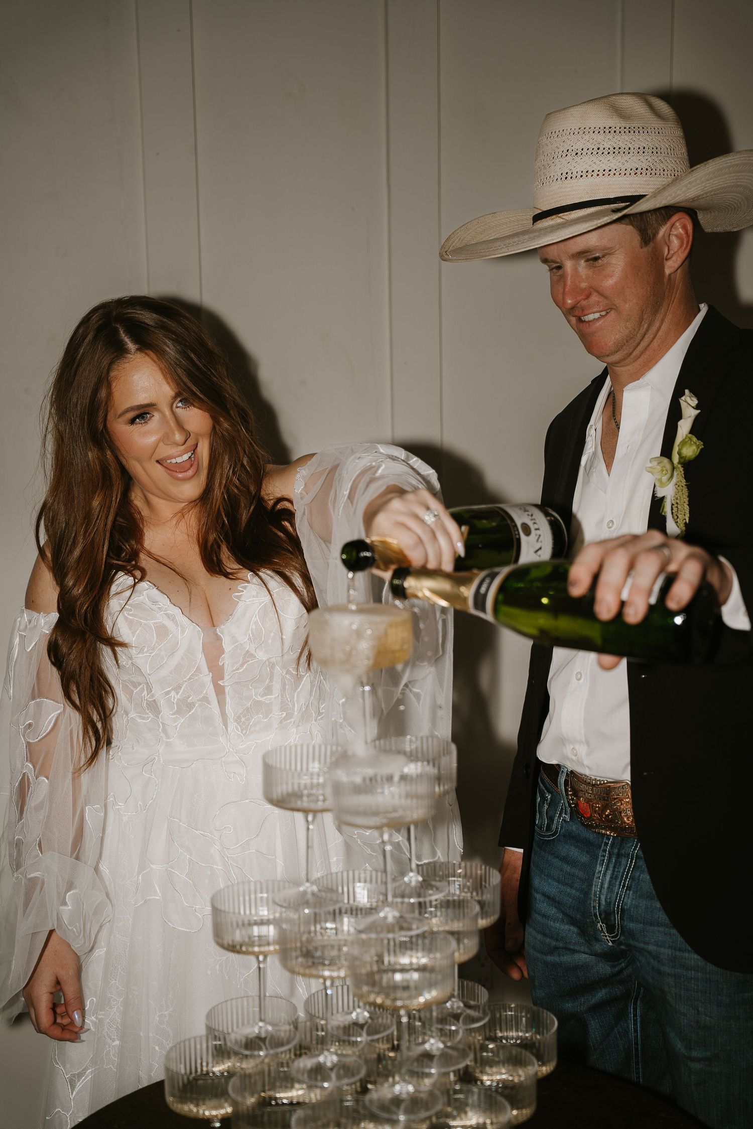 Smiling bride and groom in cowboy hat pour champagne tower at wedding reception.