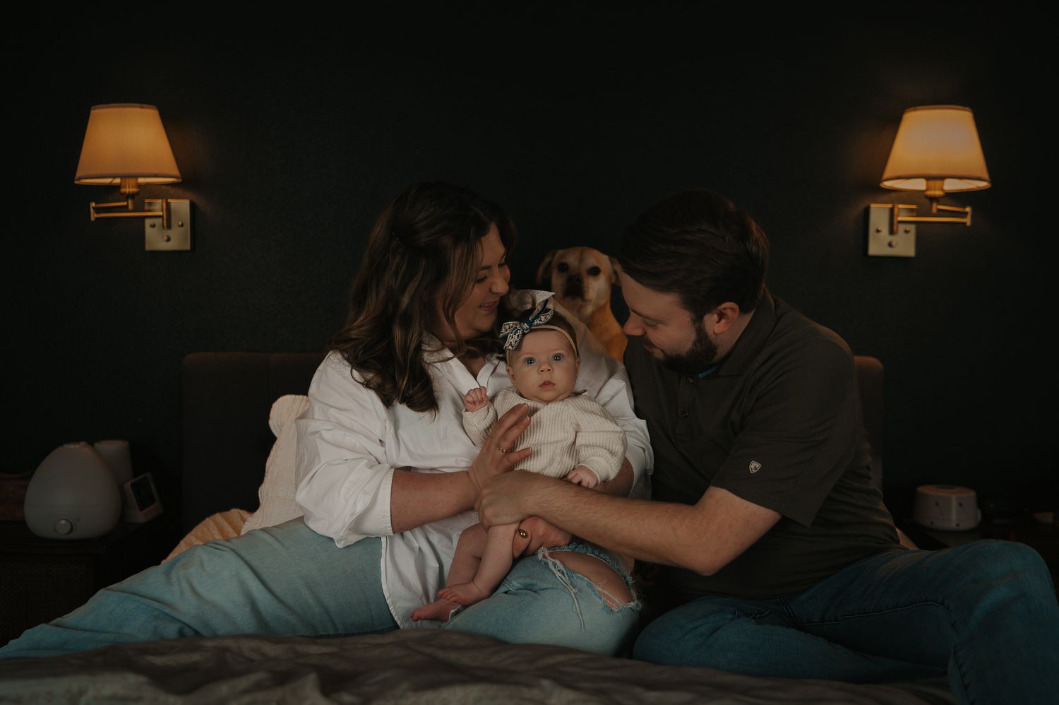 Happy parents with newborn baby sitting on bed in warmly lit bedroom.
