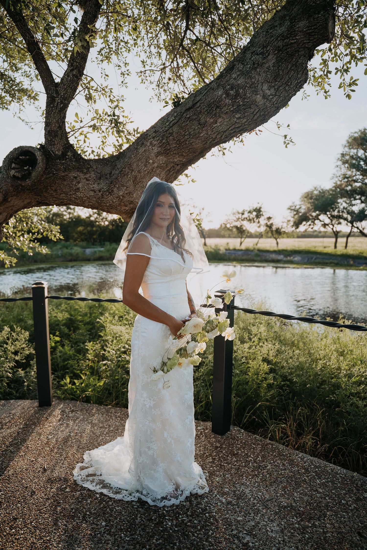Bride in lace wedding gown with veil holds bouquet beside scenic waterway under large tree.