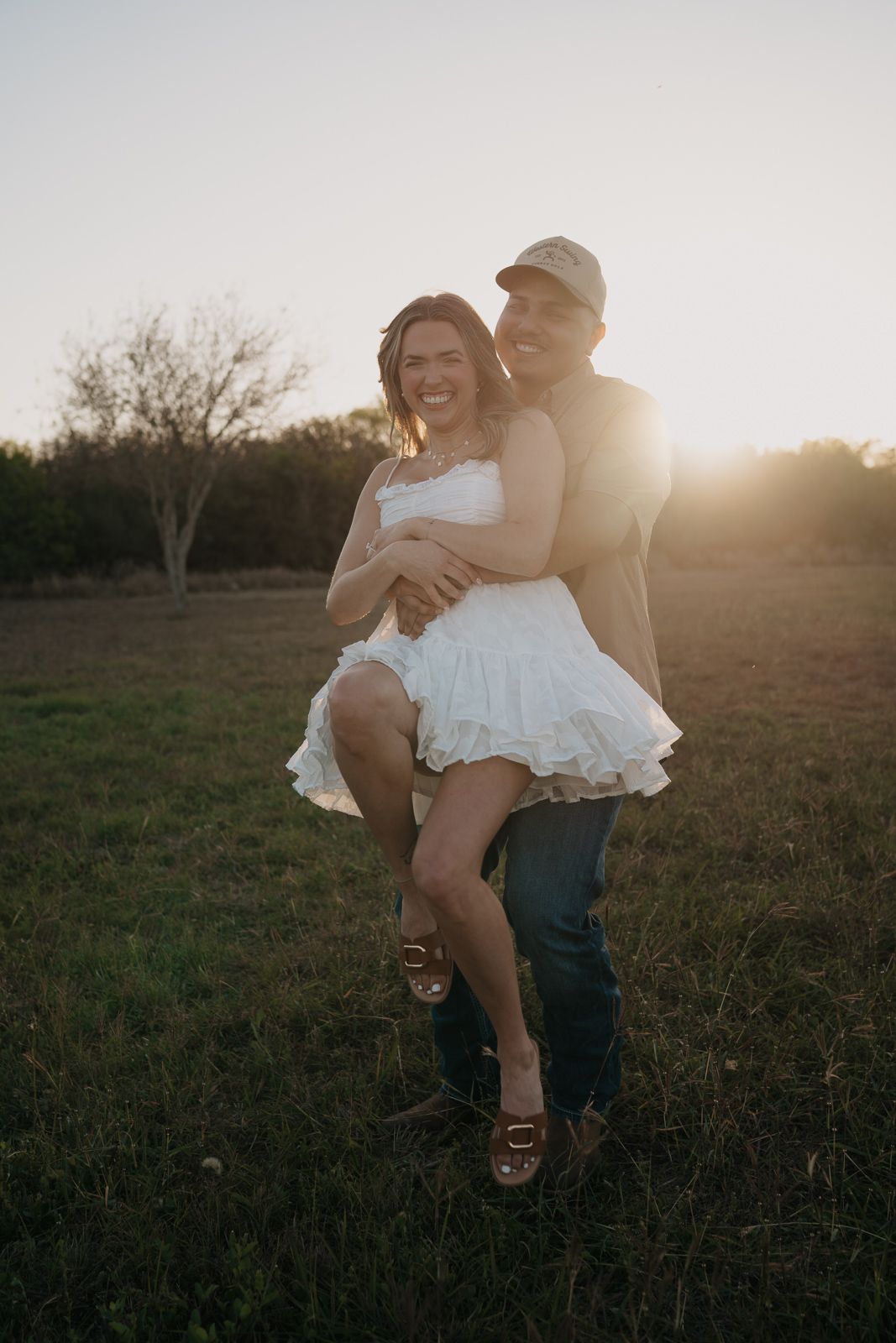 Man lifting laughing woman in white dress during a romantic sunset engagement session in a field.