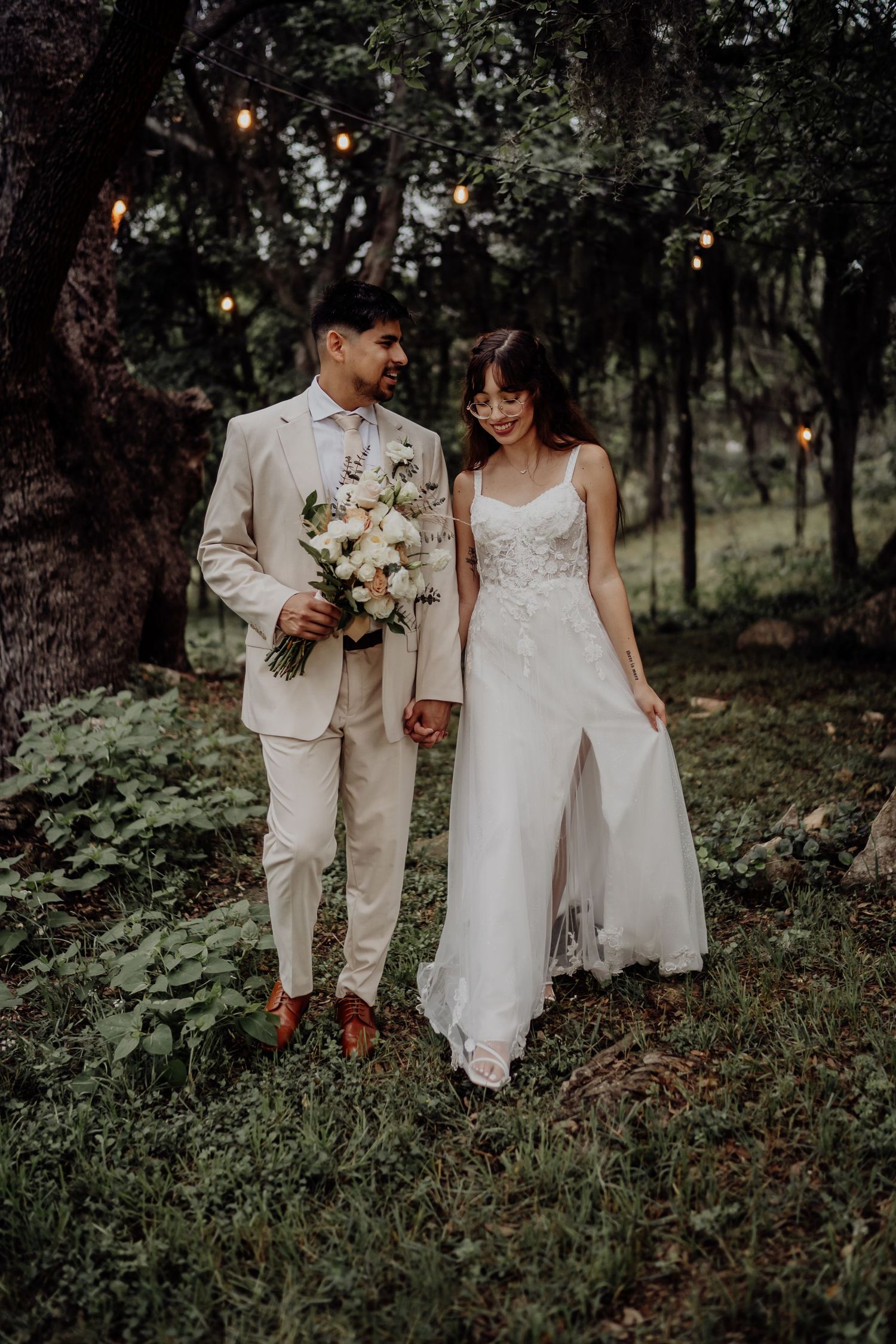 Bride and groom walking hand in hand through lush green wooded area with string lights.