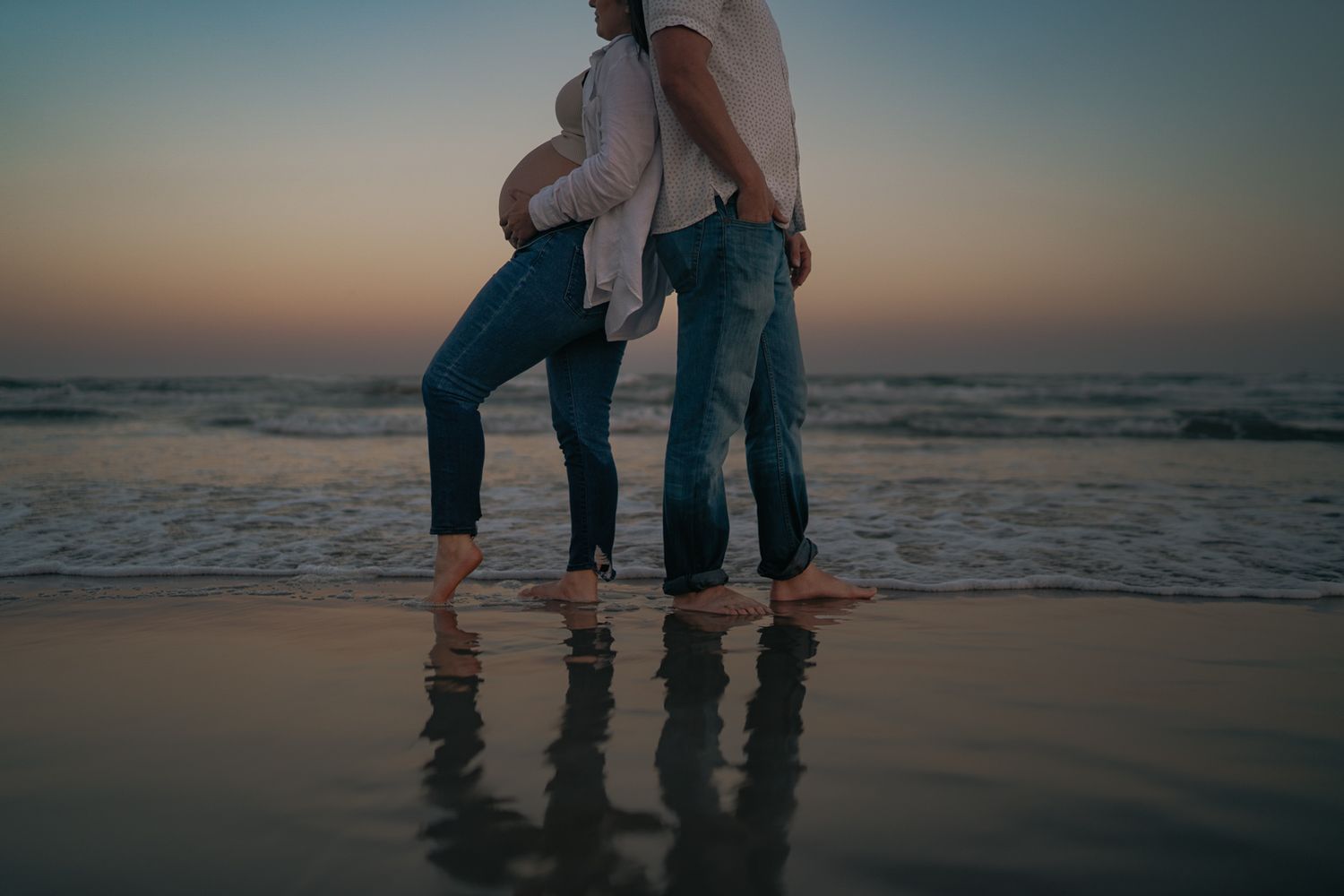 Expecting couple holding hands barefoot on ocean beach at dusk during maternity shoot.