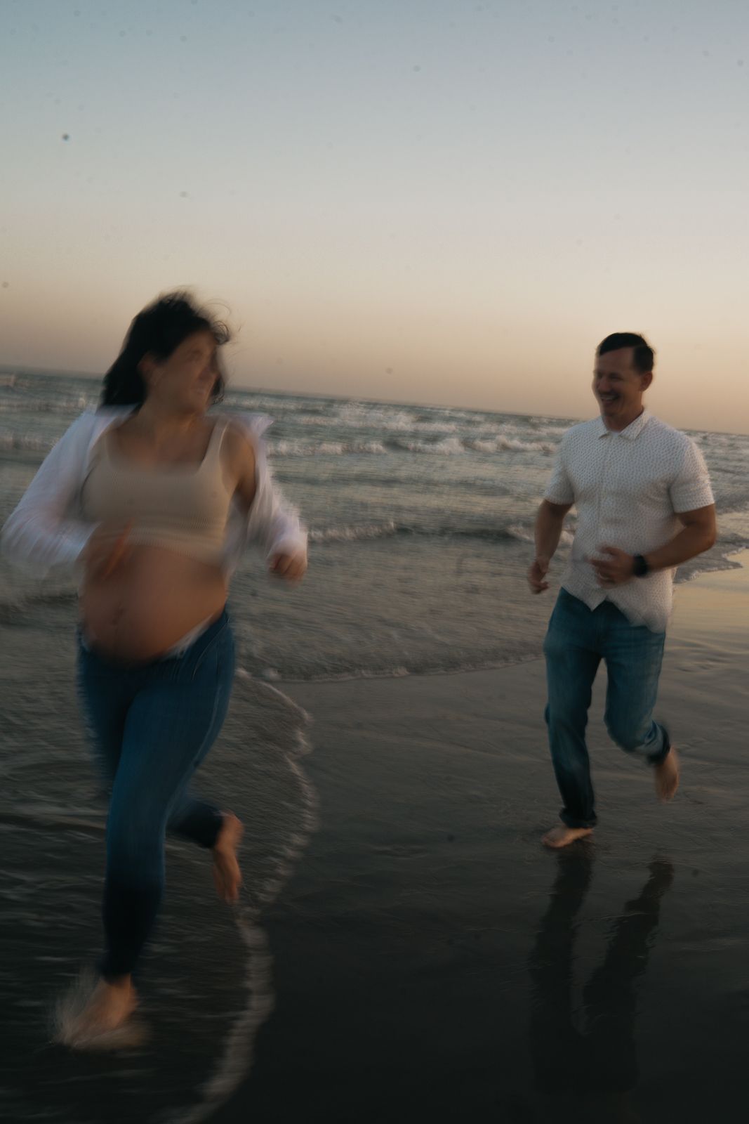 Pregnant couple running barefoot on beach at sunset.