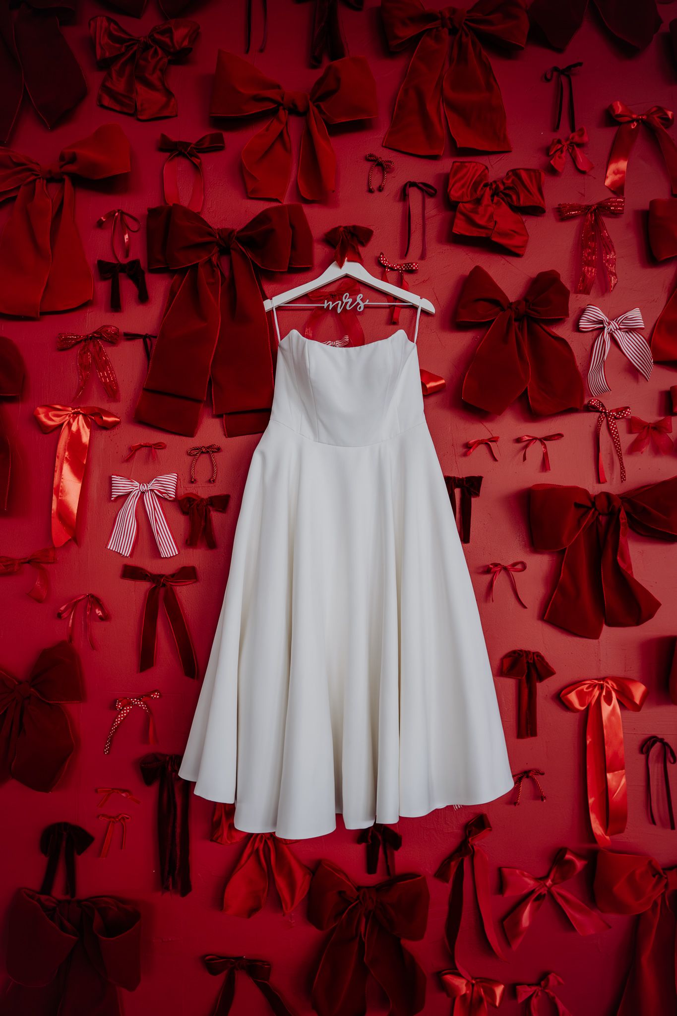 White strapless wedding dress hanging on Mrs. hanger against red wall decorated with bows.