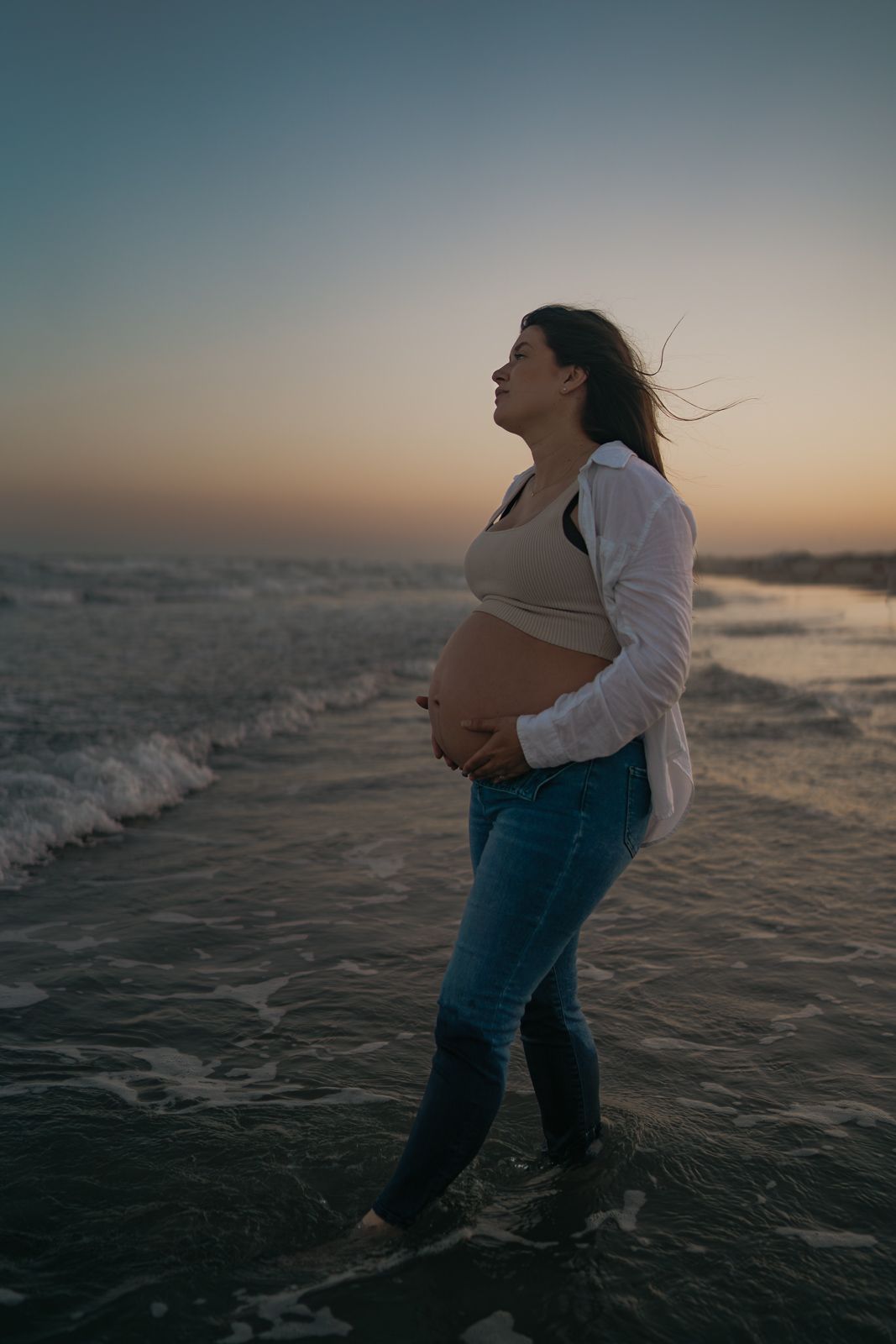 Pregnant woman cradling her bare belly while standing in ocean waves at sunset on a beach.