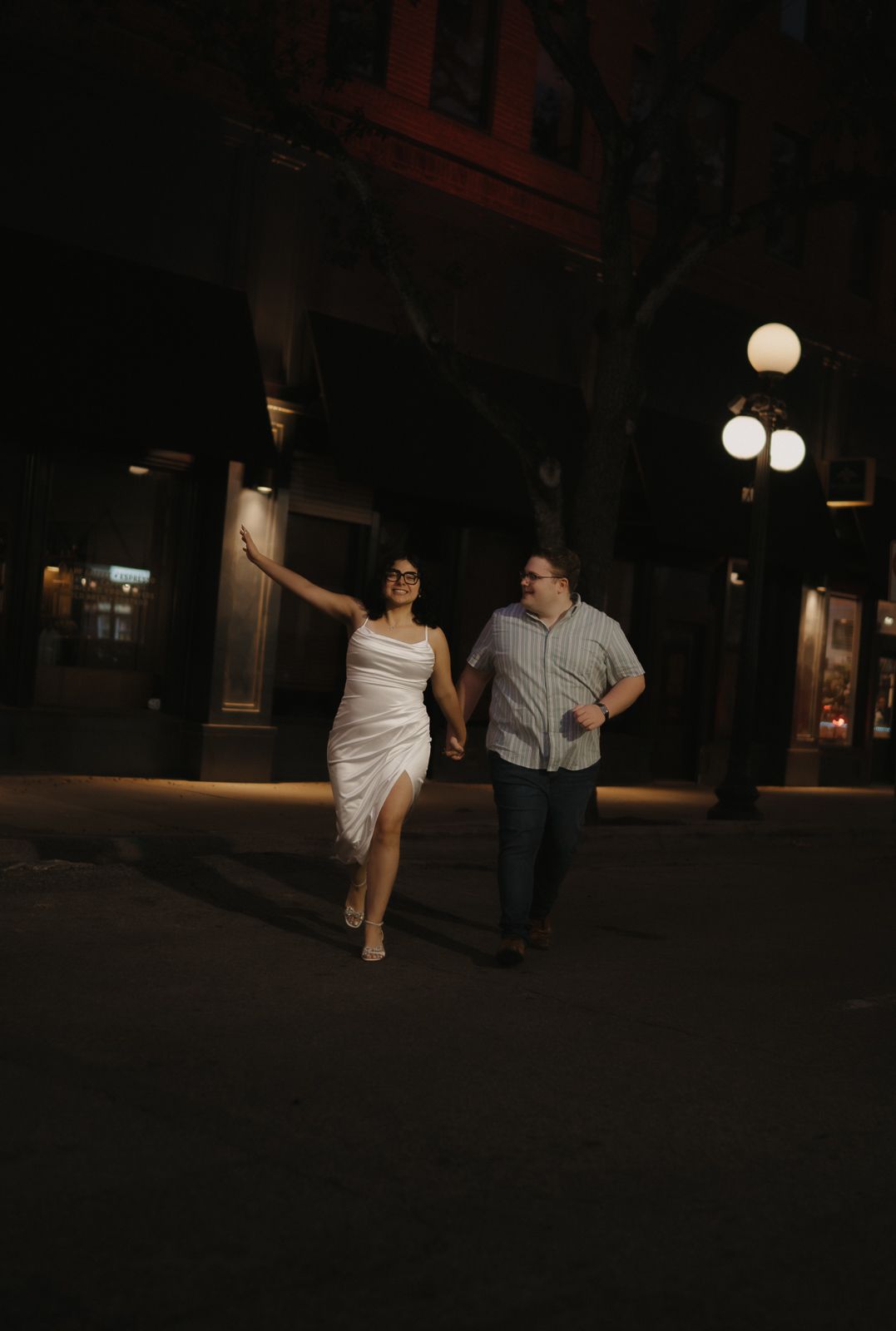 Couple walking hand-in-hand at night on a city street, woman in white dress, man in casual attire.